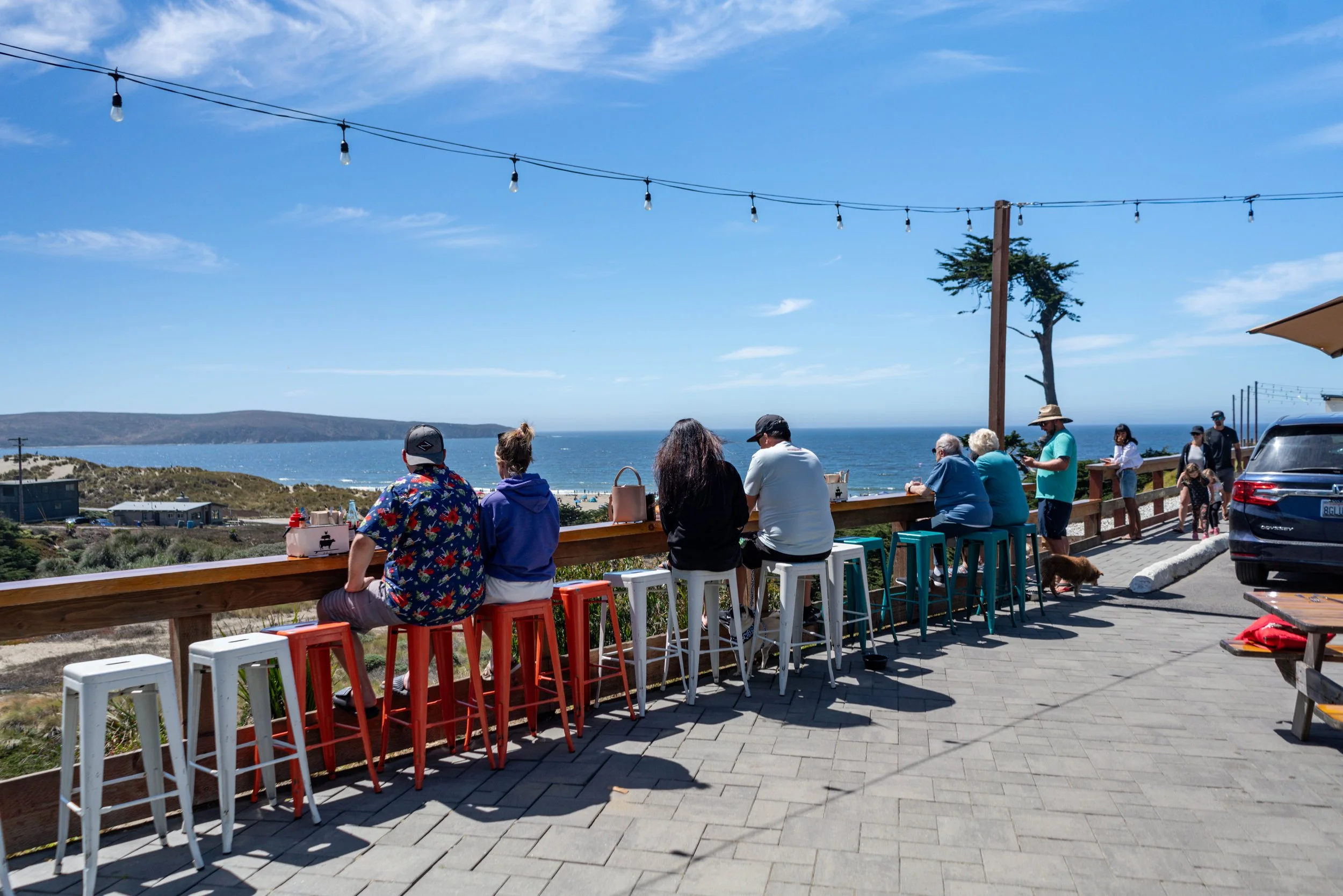 People sitting and standing at an outdoor seaside cafe with a view of the ocean and hills, under string lights and a clear blue sky.
