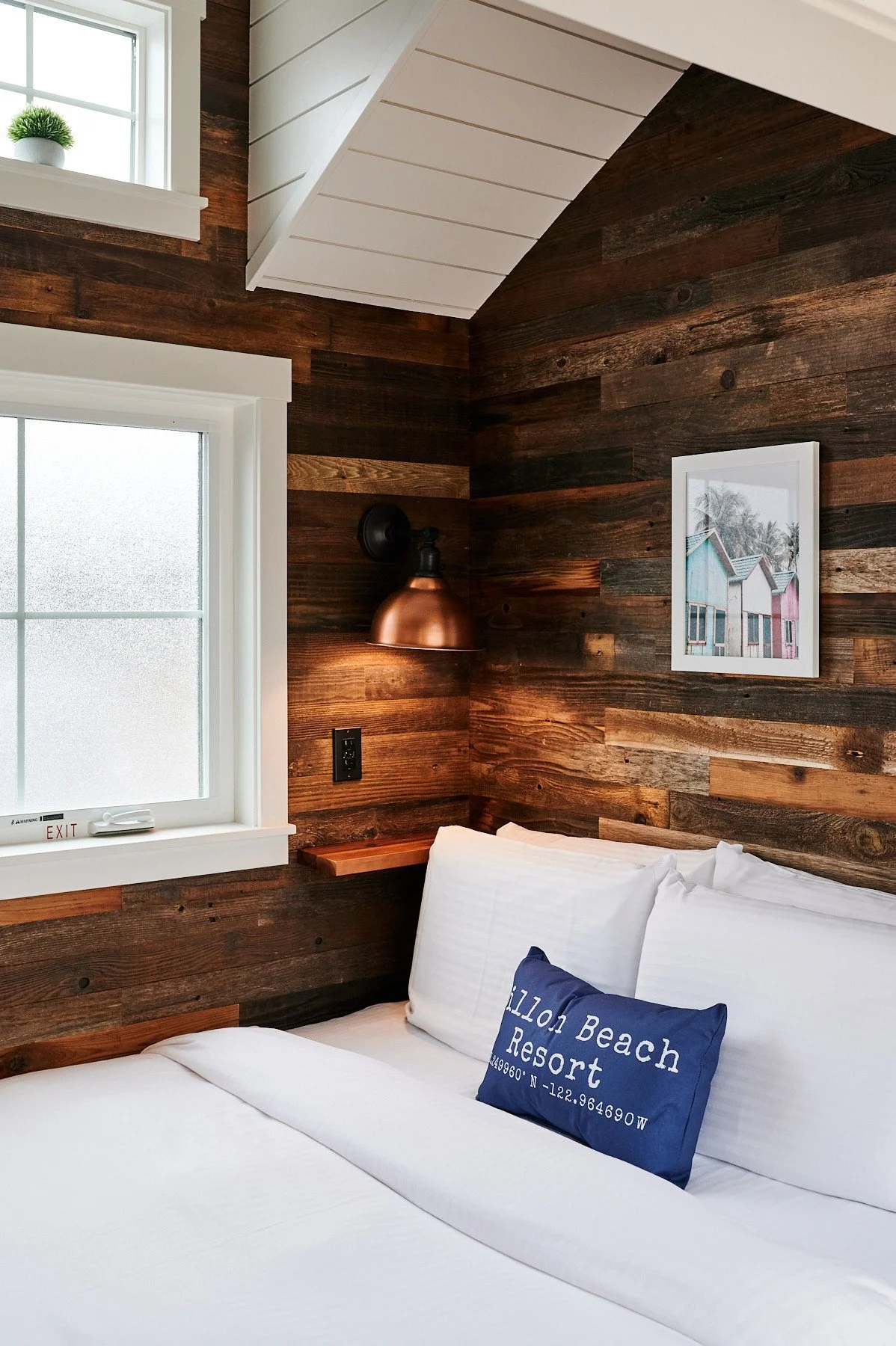 Accessible sleeping nook featuring reclaimed dark wood plank walls, a copper bedside sconce, and white linens with a Dillon Beach Resort accent pillow.