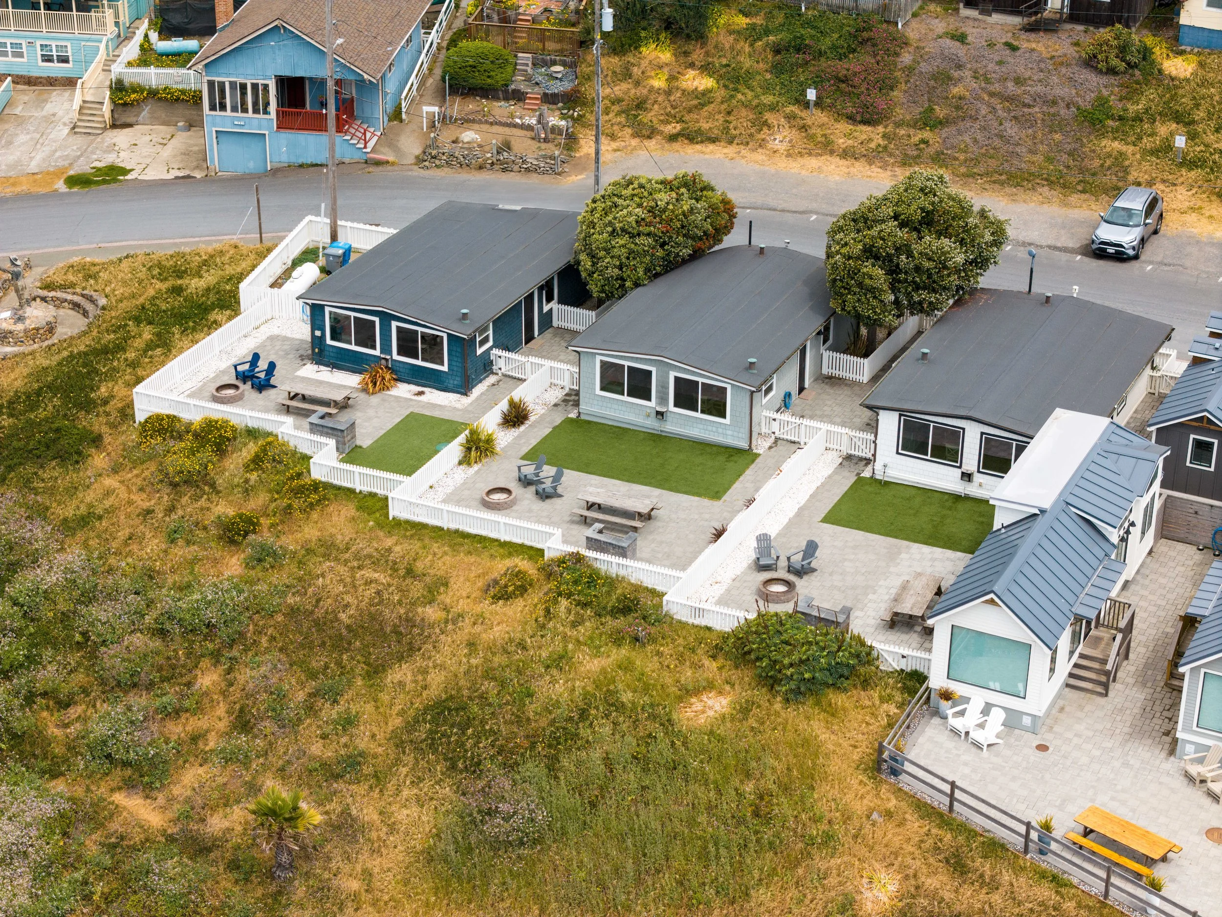 Aerial view of multiple coastal cottages at Dillon Beach Resort, each with white picket fences, private gravel patios, fire pits, and artificial grass lawns, all overlooking the grassy coastal dunes.