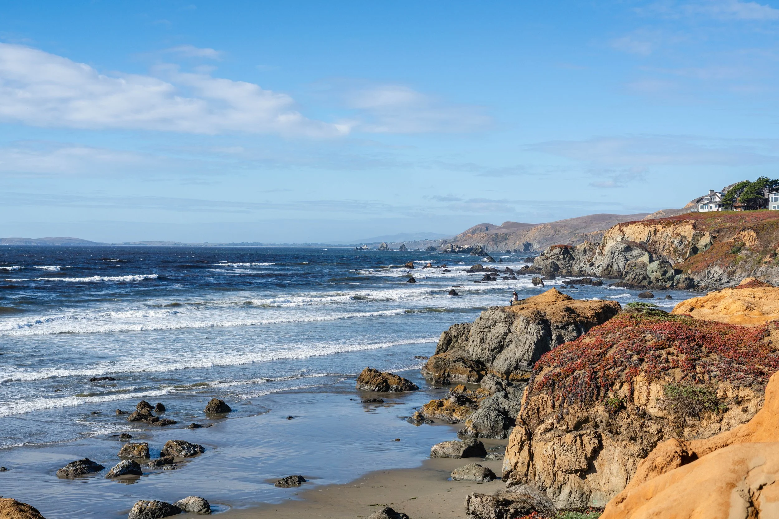Coastal scene with rocky shoreline, waves, and a house on a hill.