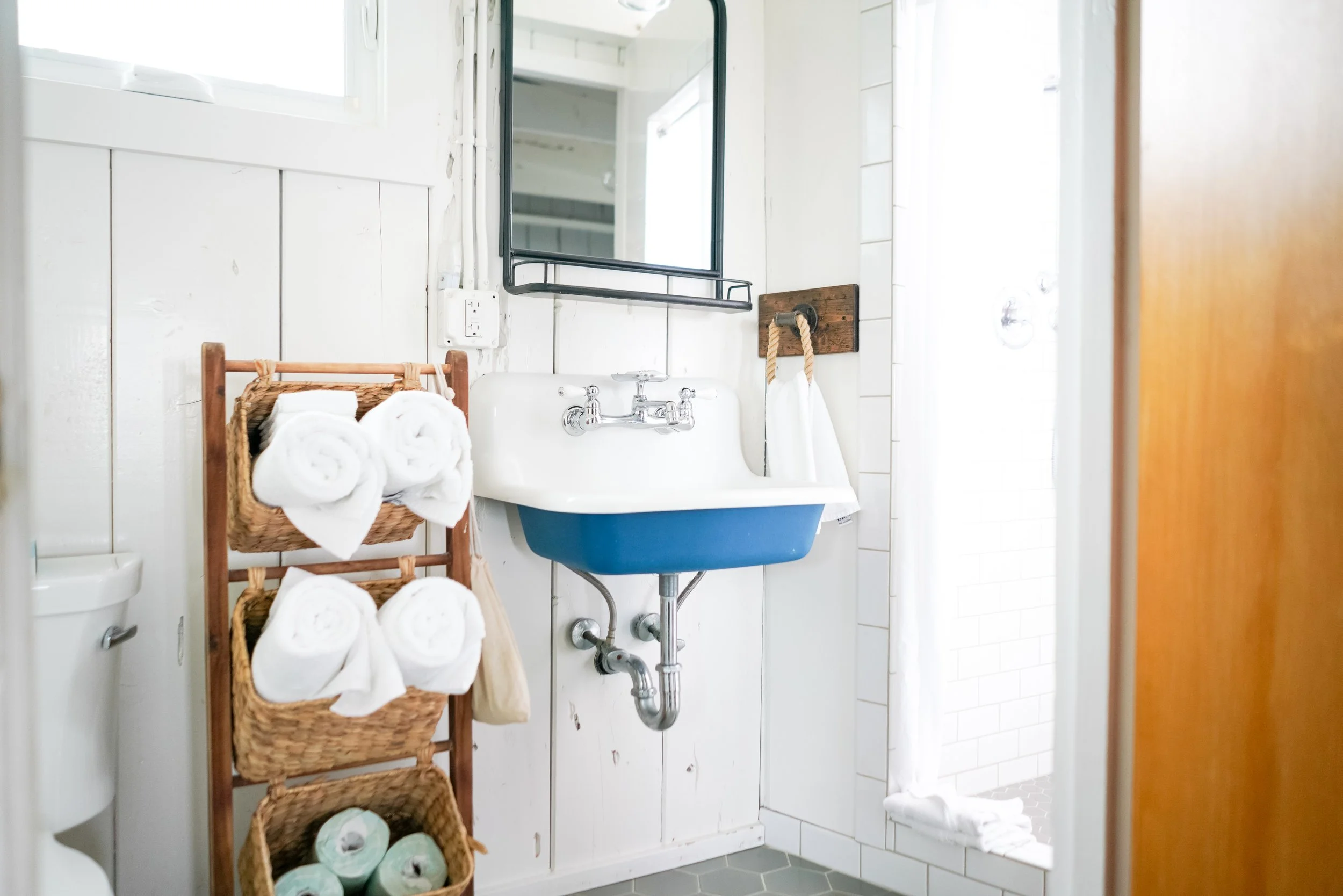 Bright bathroom with a white and blue vintage wall-mount sink, white shiplap walls, and a wooden towel rack with wicker storage baskets.