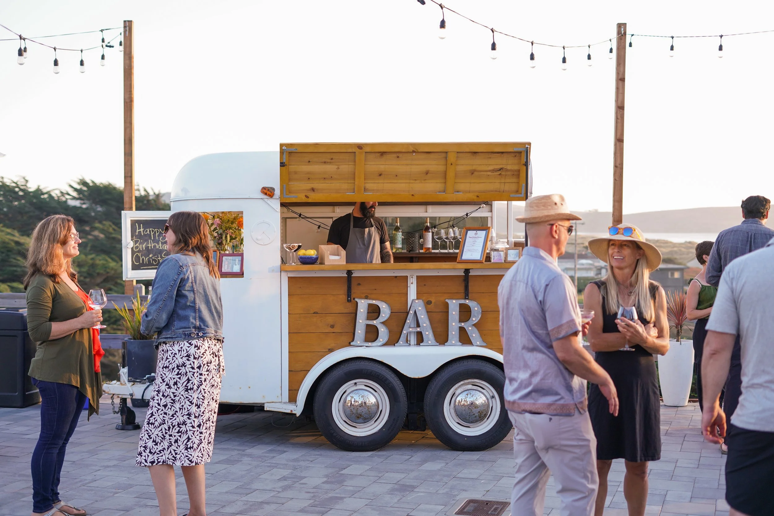 An outdoor evening event at Dillon Beach Resort, featuring a unique mobile bar trailer with a marquee 'BAR' sign, string lights, and guests mingling.