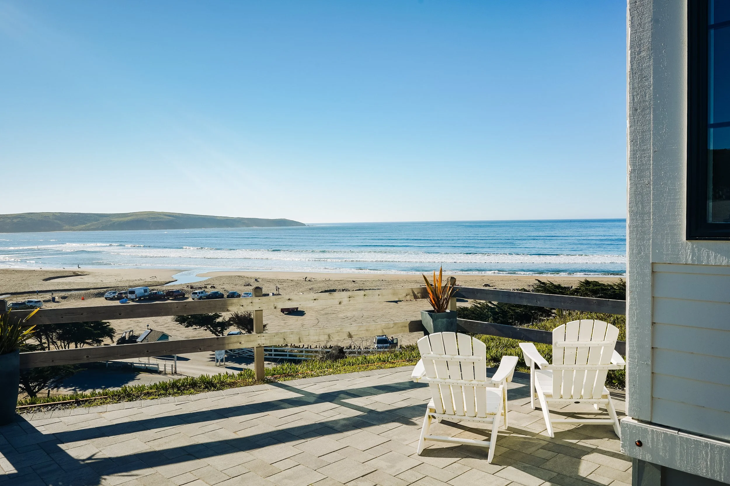 Two white Adirondack chairs on a stone patio overlooking the ocean and sandy beach at Dillon Beach Resort on a clear, sunny day.
