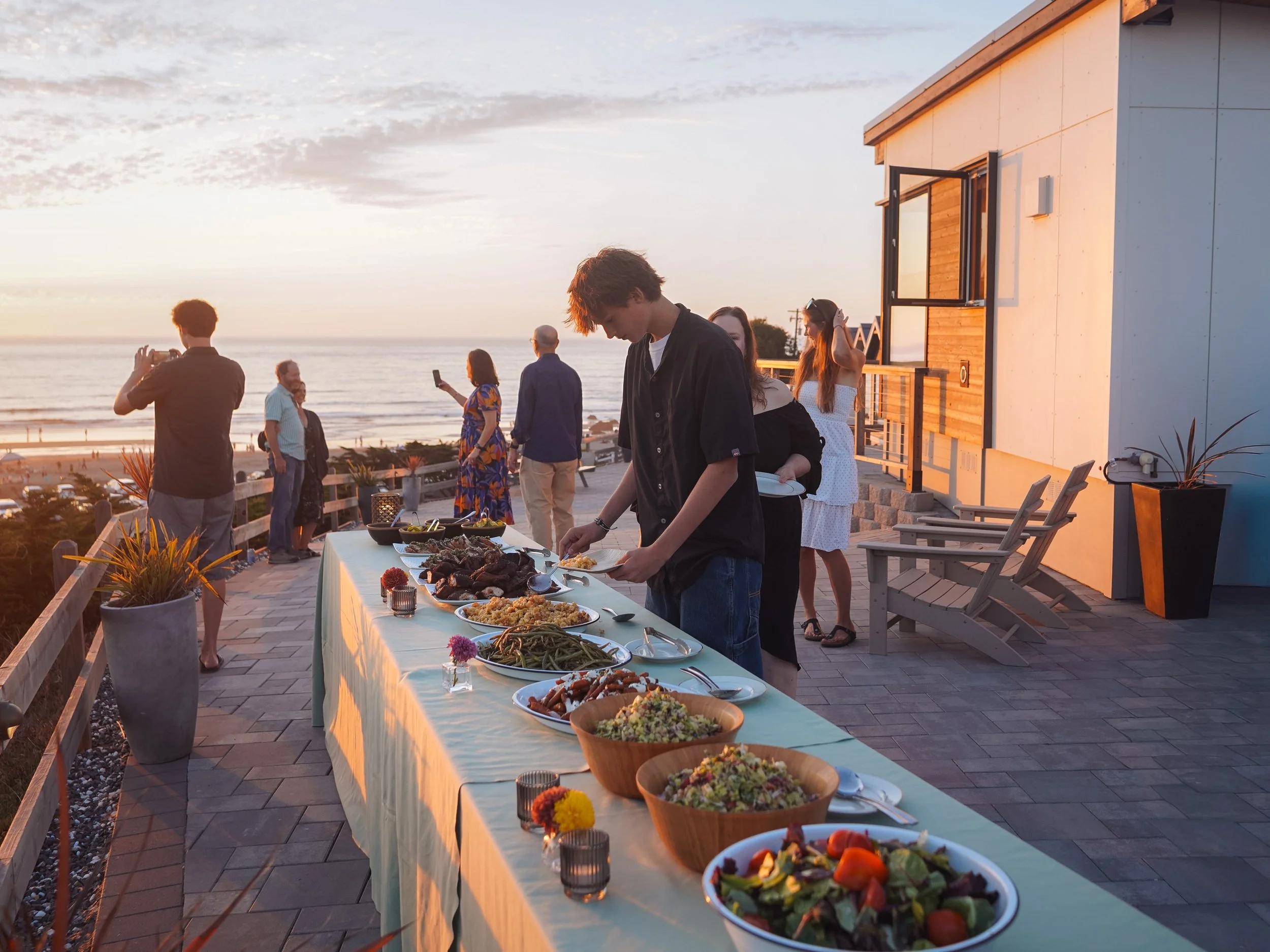 People enjoying an outdoor buffet by the beach at sunset, with various dishes on a long table, and ocean in the background.