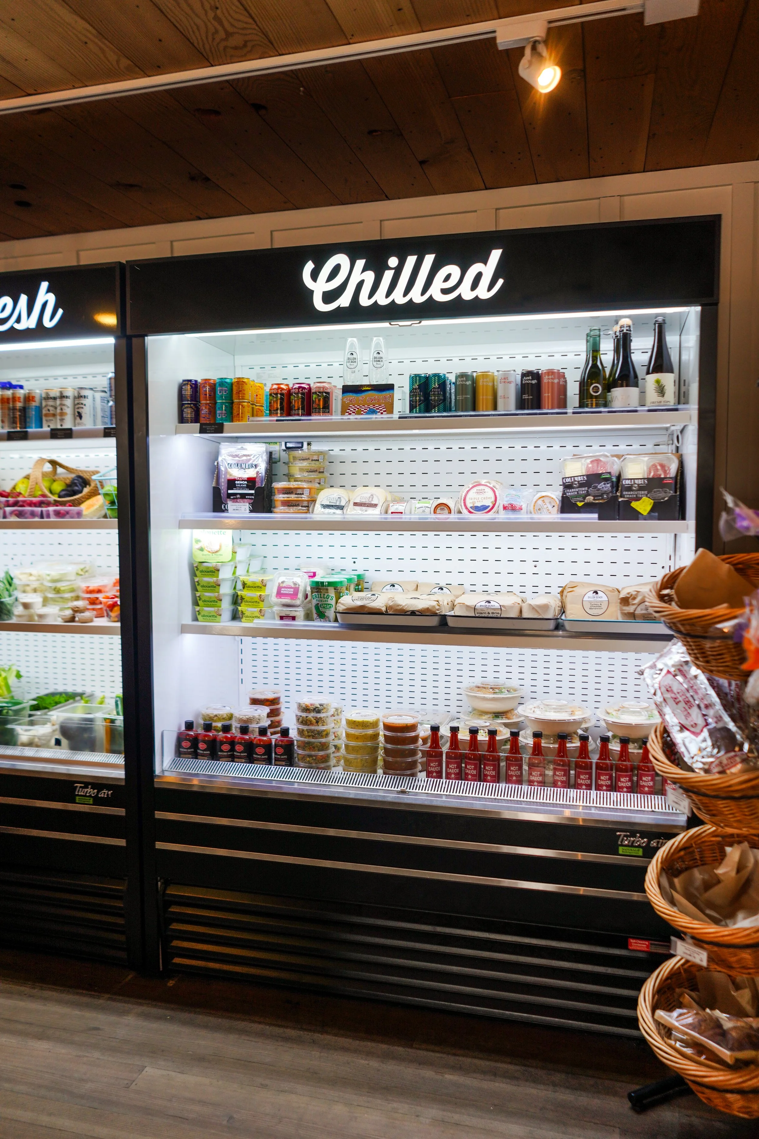 Refrigerated grocery display case labeled 'Chilled' with various beverages, dairy, and condiments inside, in a store aisle with wooden ceiling and woven basket with items on the side.