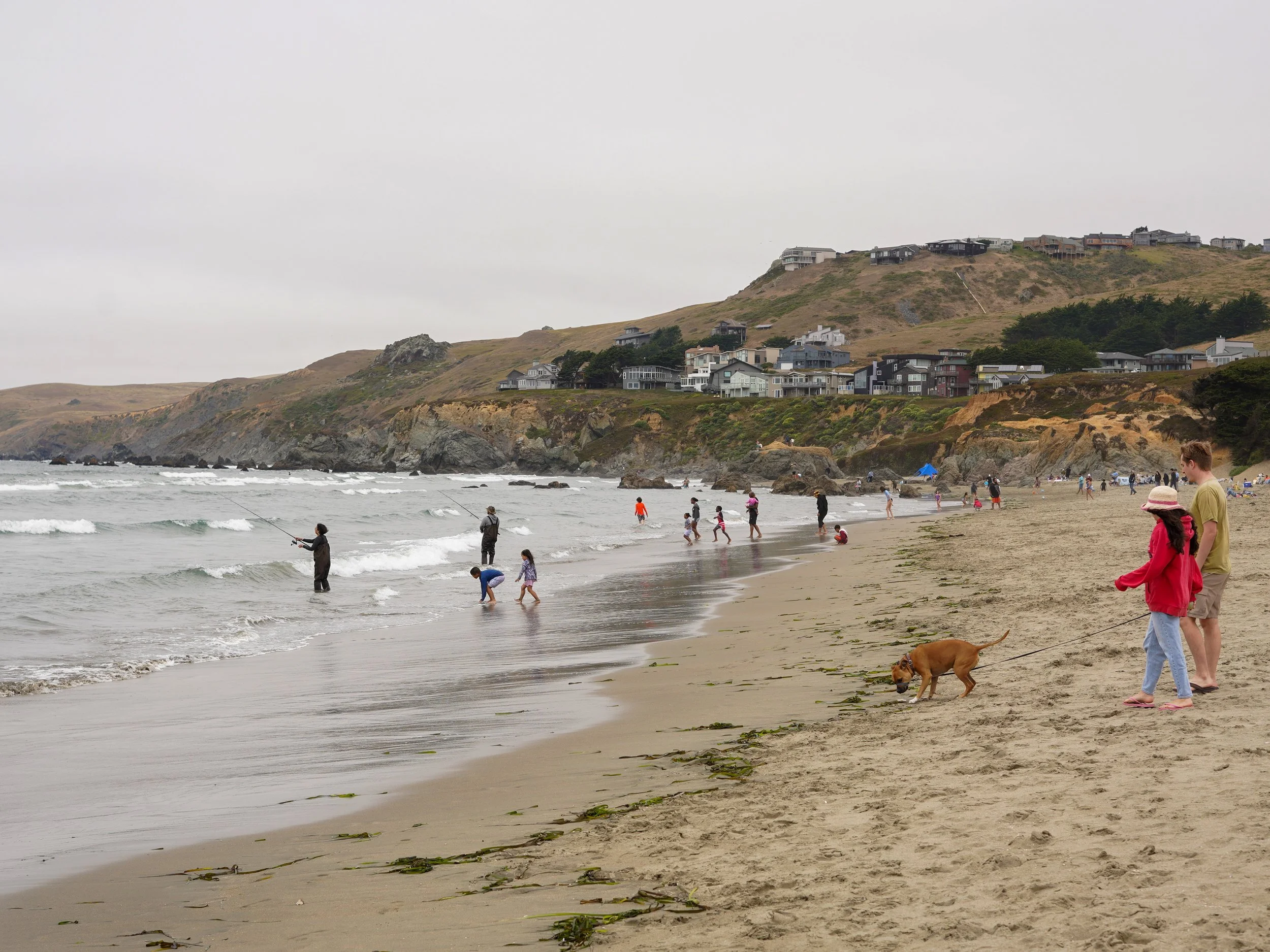 People walking and fishing on a beach with rocky cliffs and houses on a hillside in the background.