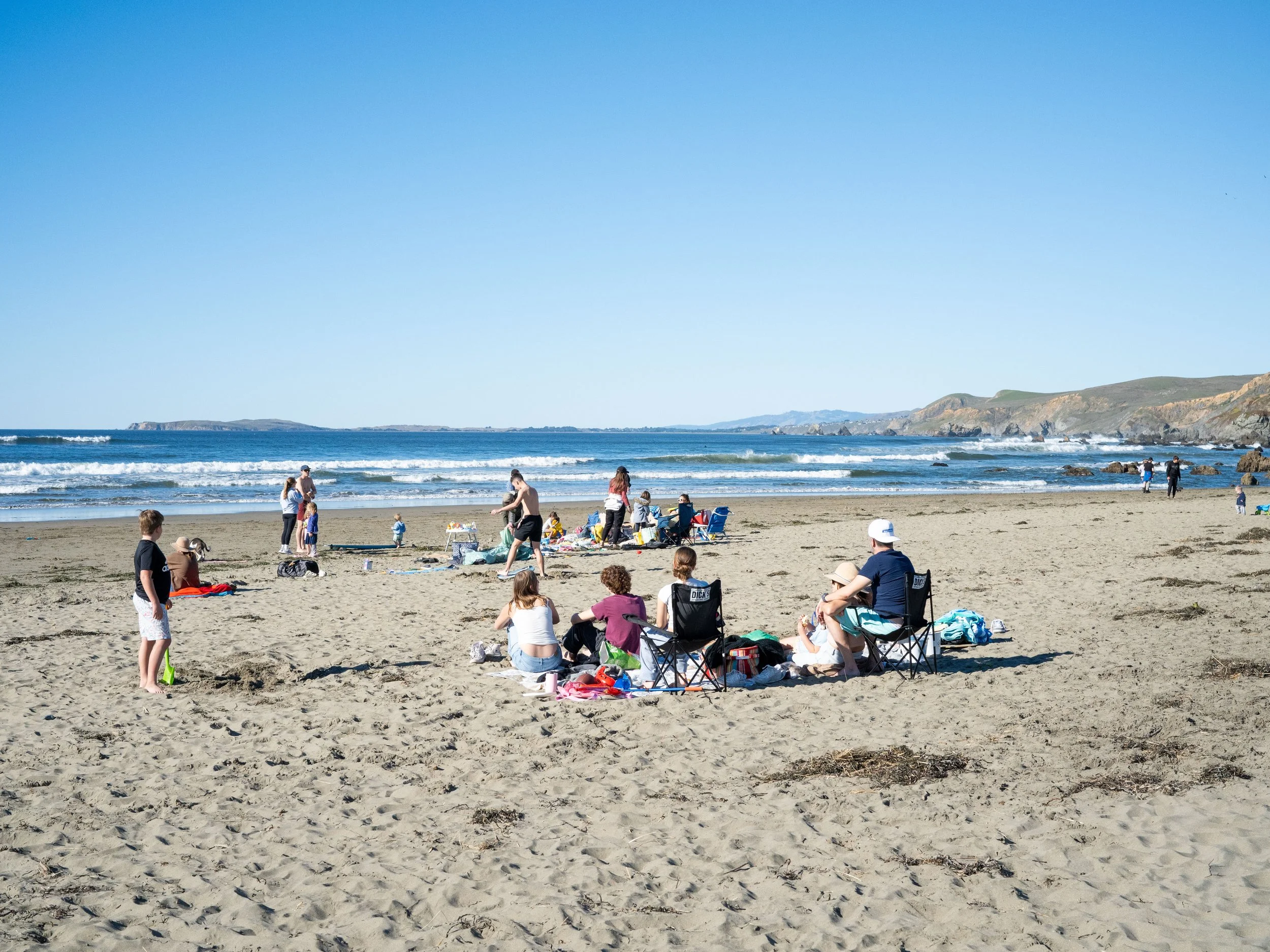 People enjoying a sunny day at the beach with clear blue sky, waves, and distant cliffs.