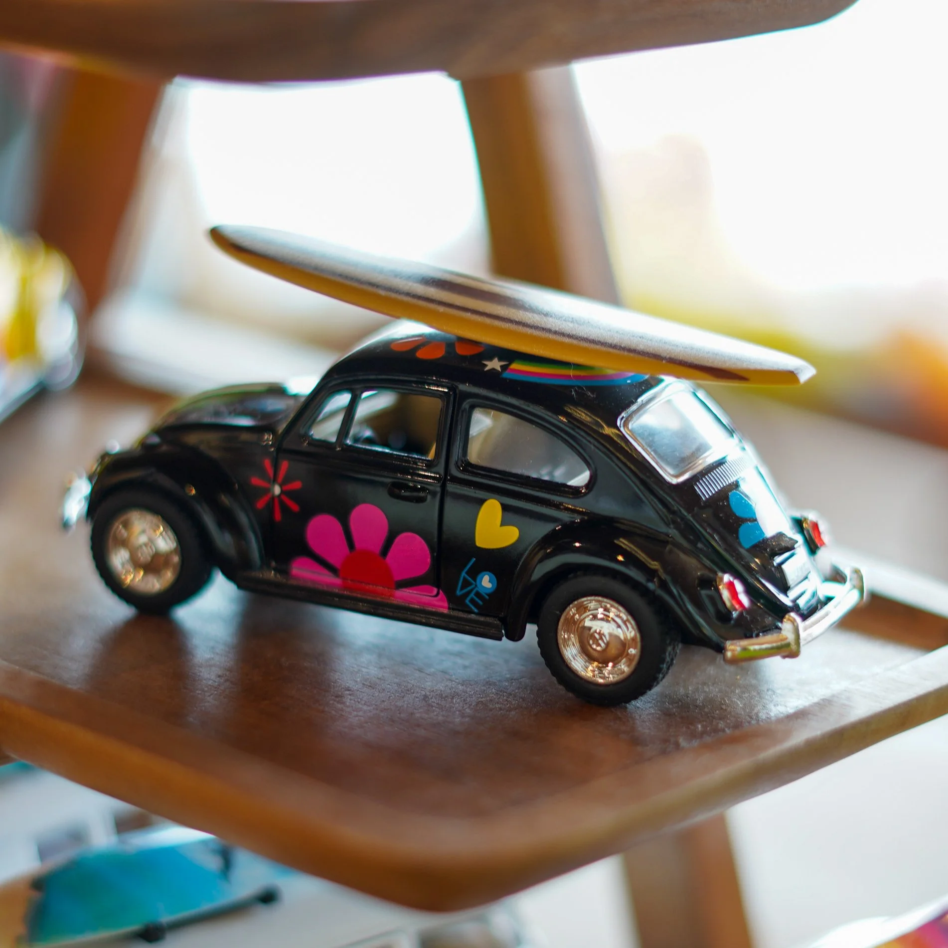 Close-up of a toy car with colorful flower and heart decals, stacked beneath a wooden furniture piece, with a skateboard and other objects in the background.