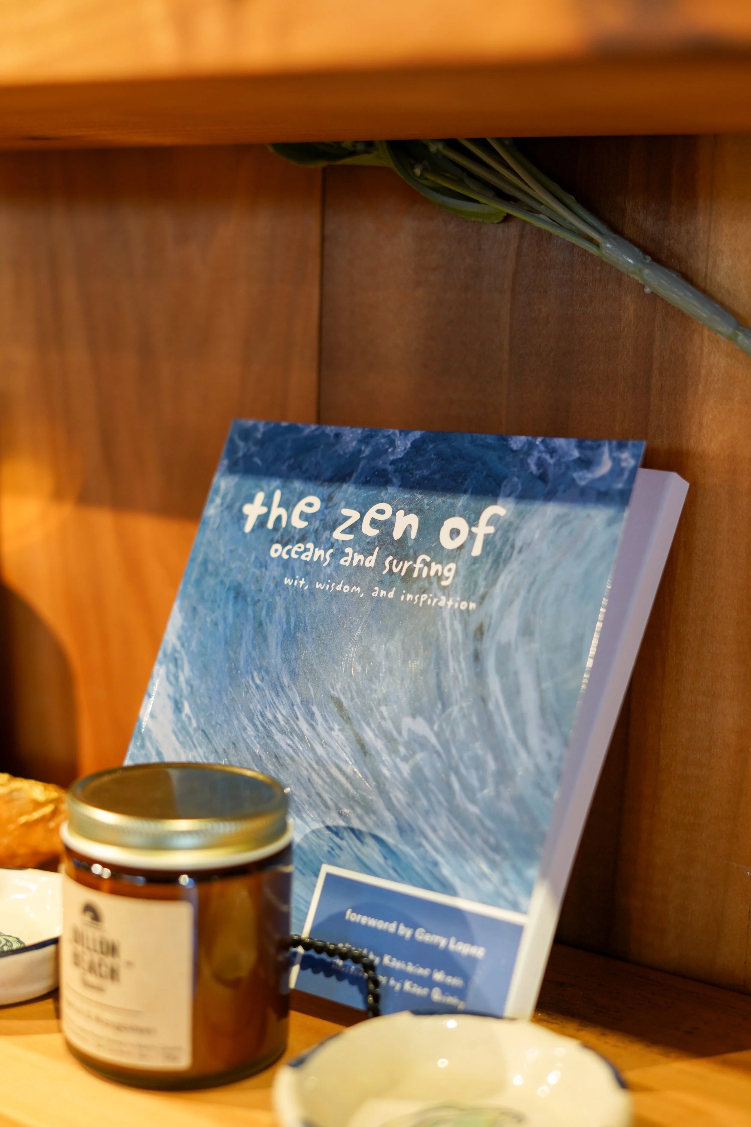Close-up of a bookshelf with a book titled "the zen of oceans and surfing" and various items including a jar and a bowl.