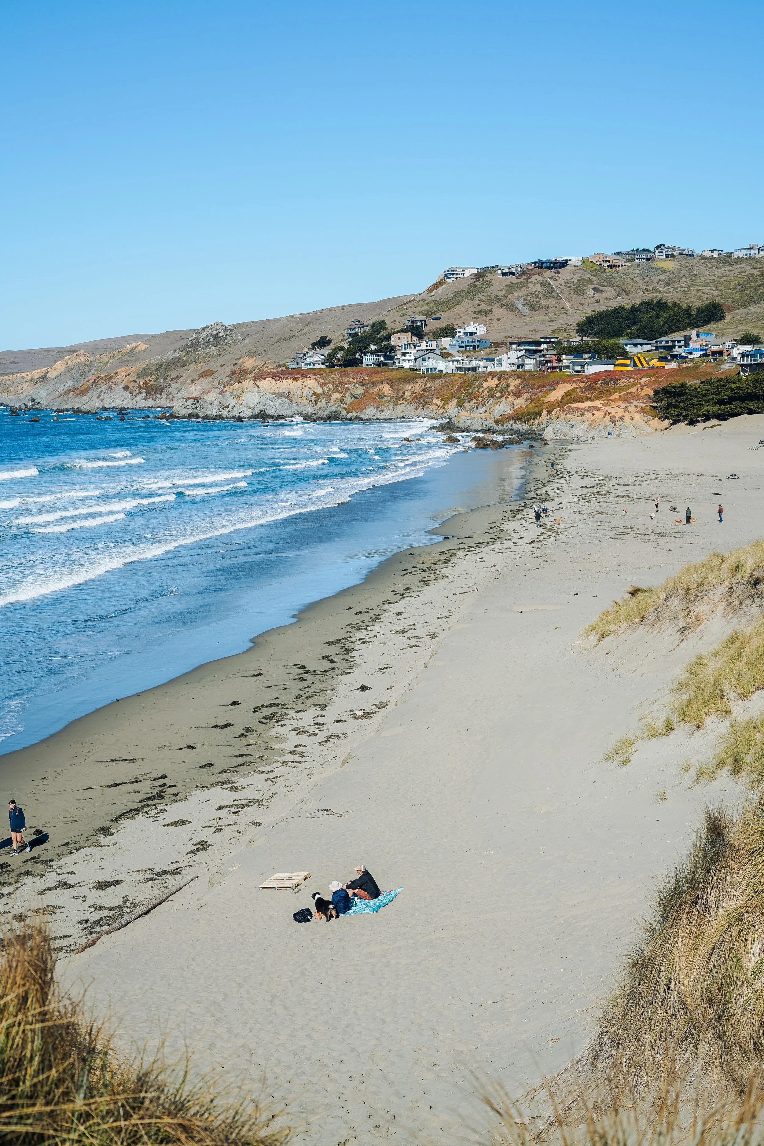 People relaxing on a sandy beach with cliffs and houses in the background, and the ocean waves crashing onto the shore under a clear blue sky.