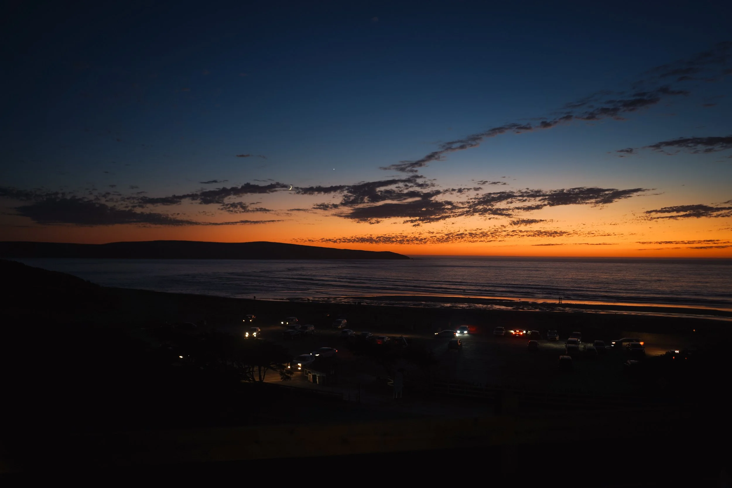 Sunset over the ocean with a dark shoreline, scattered clouds in the sky, and cars parked along a road in the foreground.