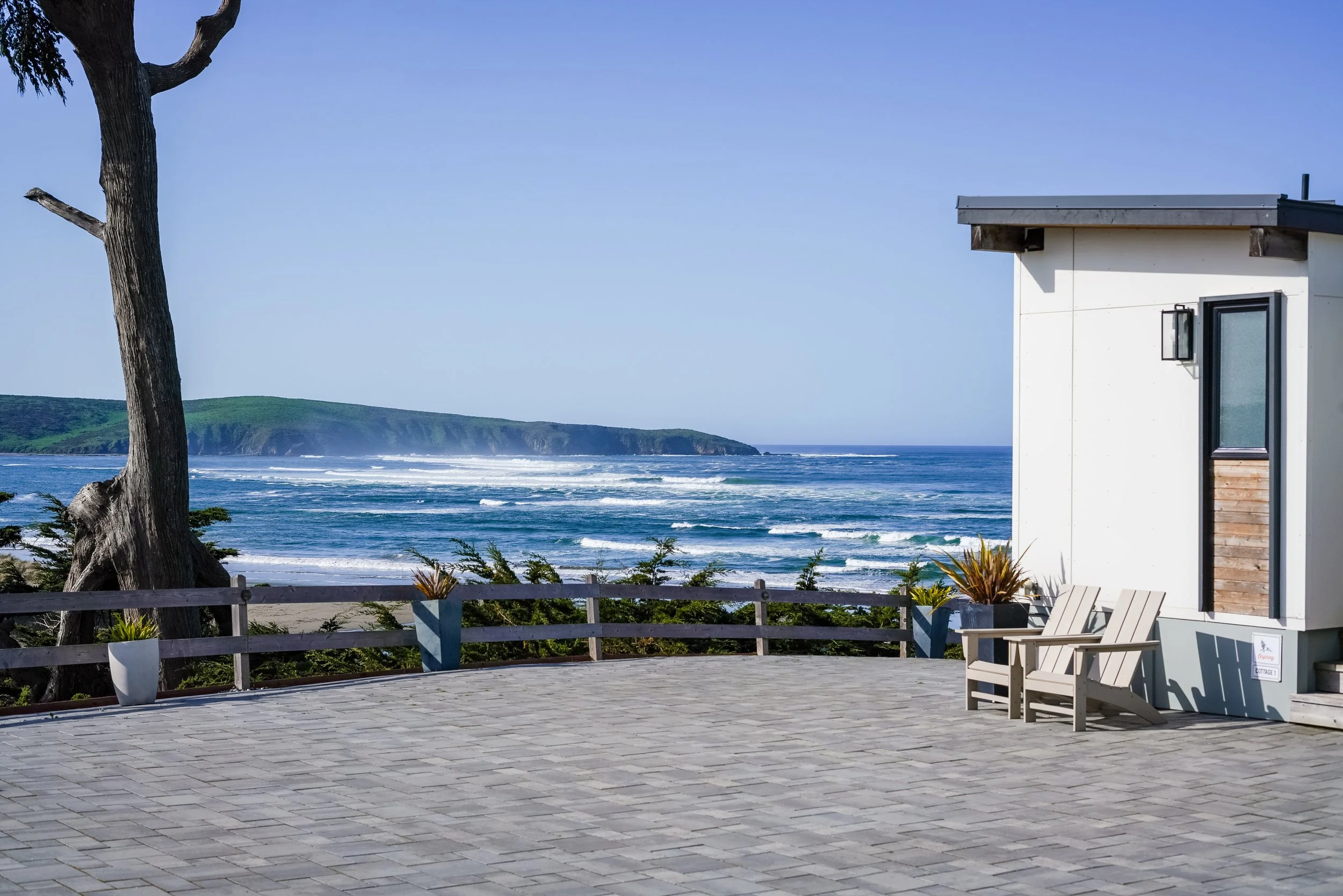 A spacious stone event patio at Dillon Beach Resort featuring a modern white cabin, Adirondack chairs, and an unobstructed panoramic view of the Pacific Ocean.