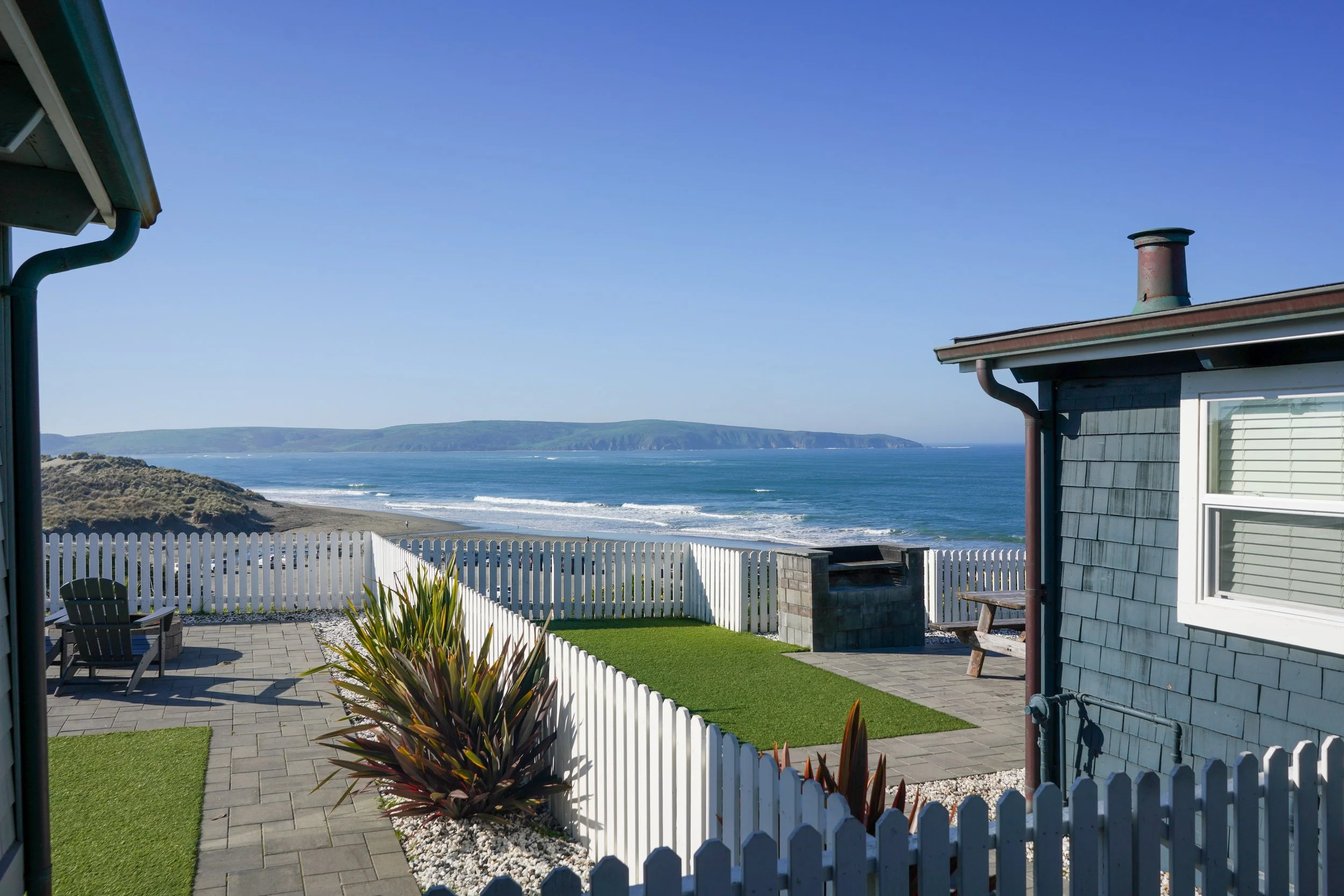 Looking between two cedar-shingle beach houses toward a white picket fence and a panoramic view of the California coastline and ocean waves.