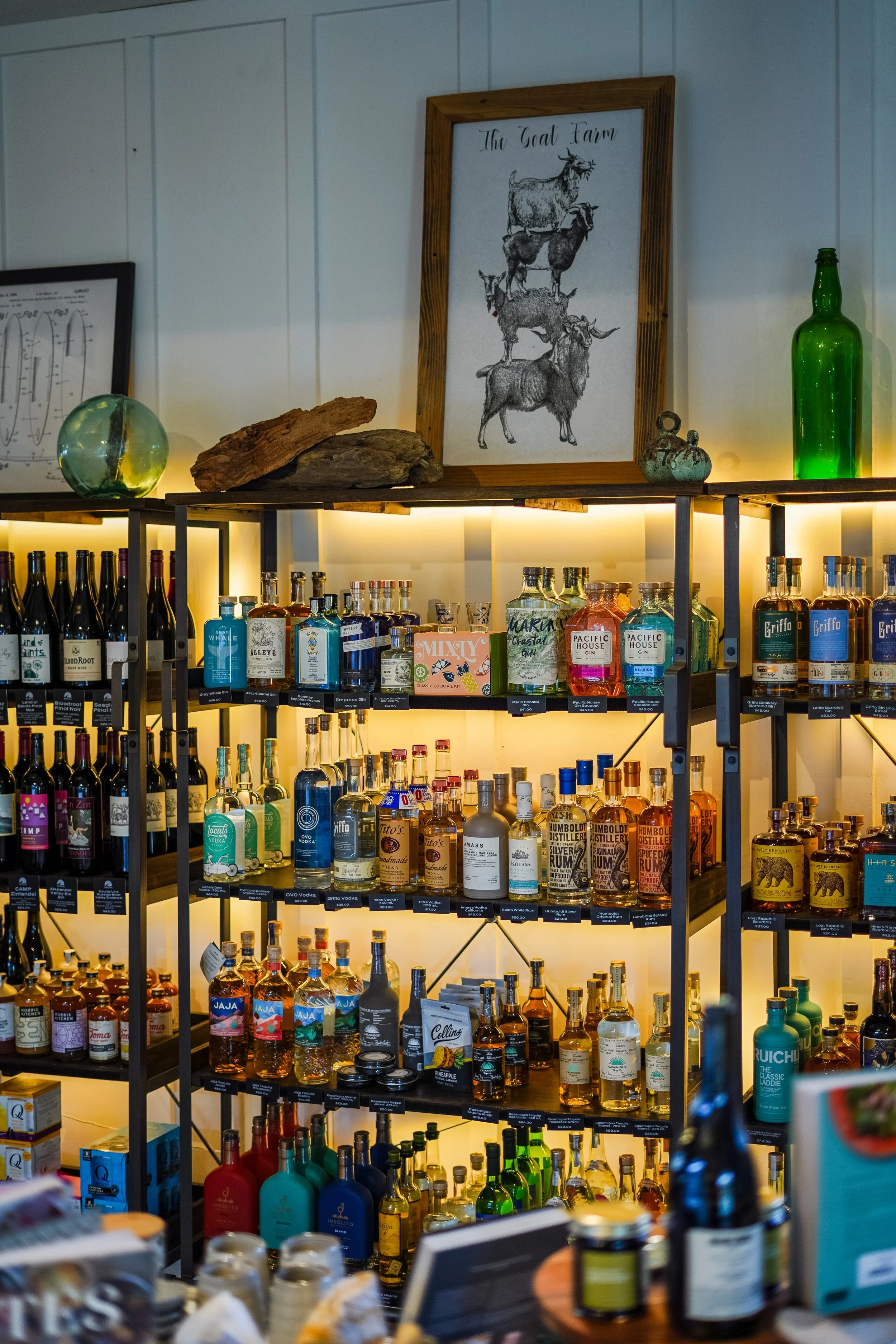 A bar display with liquor bottles, framed artwork of goats titled 'The Goat Farm,' decorative items, and a green glass bottle on a wooden shelf with warm lighting.
