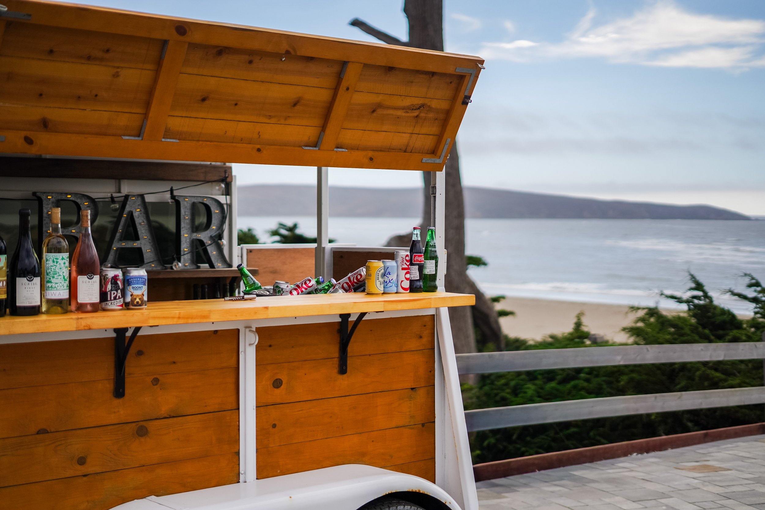 A wooden mobile bar cart outdoors with various bottles of alcohol and soda cans, with a backdrop of a lakeside view and cloudy sky.
