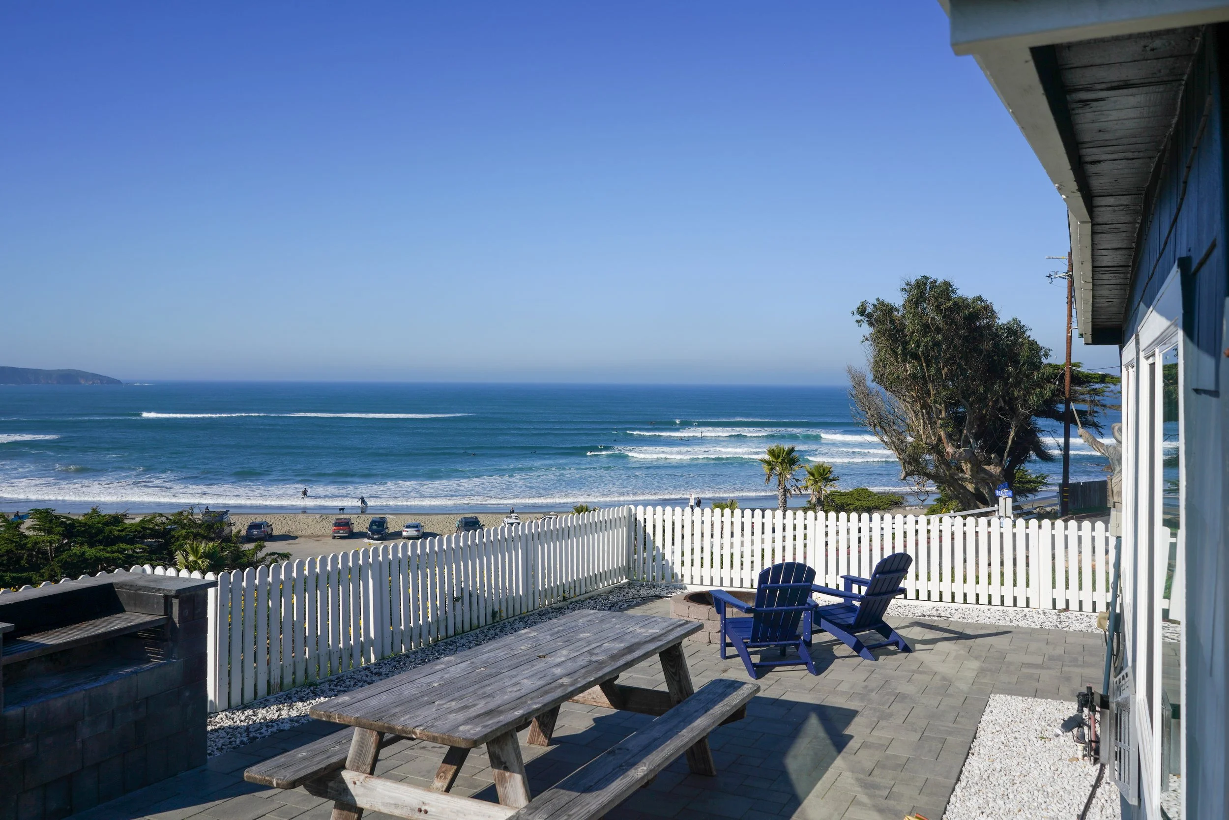 Panoramic view from a stone patio with a white picket fence, featuring a wooden picnic table and blue Adirondack chairs looking out over rolling ocean waves and a sandy beach.