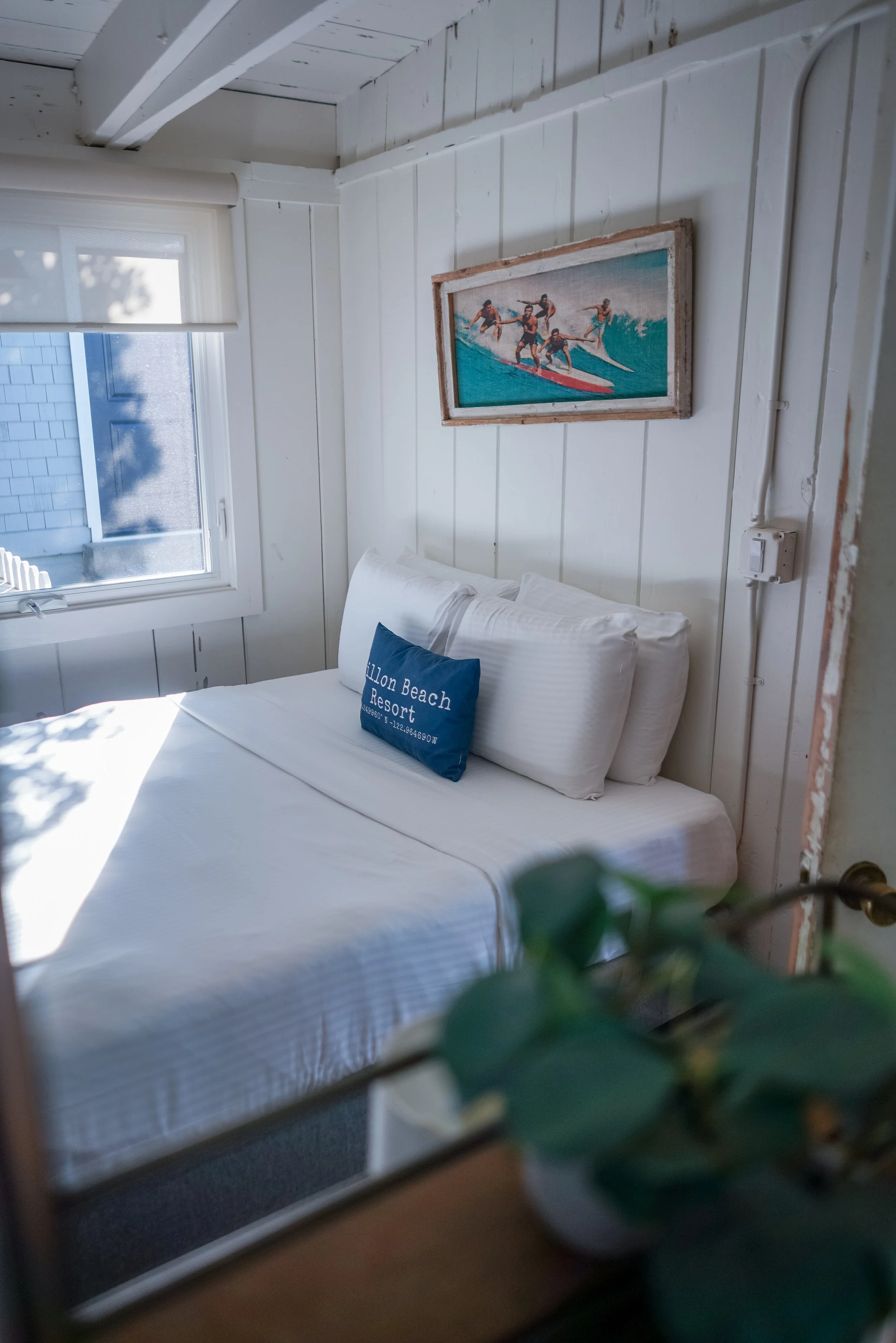 A cozy bedroom with white vertical shiplap walls, a bed with crisp white linens, a navy blue decorative pillow, and vintage-style surf art hanging above the headboard.