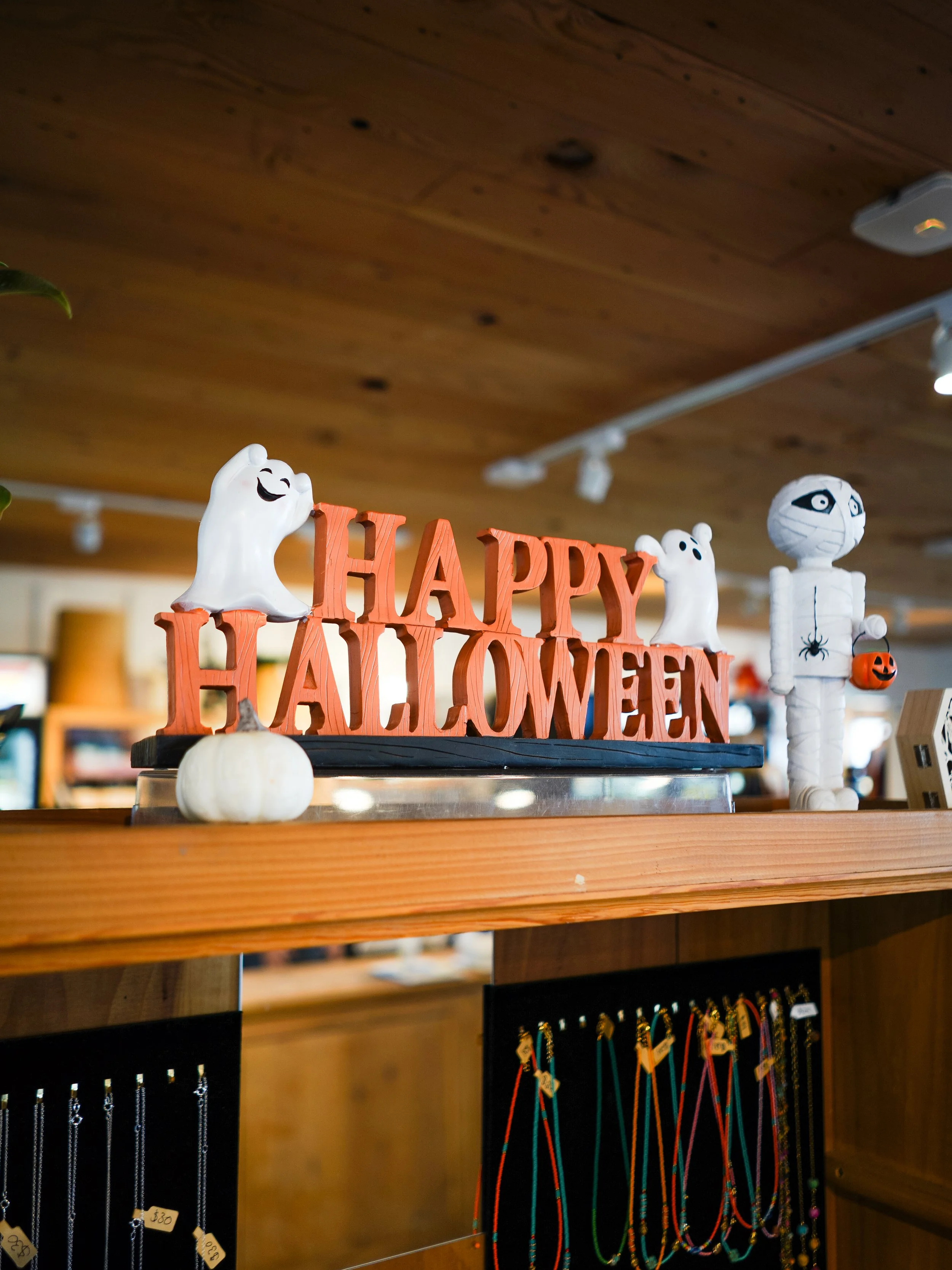 Halloween decorations including white ghost figures, a sign that says 'Happy Halloween', a mummy figure, mini pumpkins, and colorful necklaces hanging on a display.