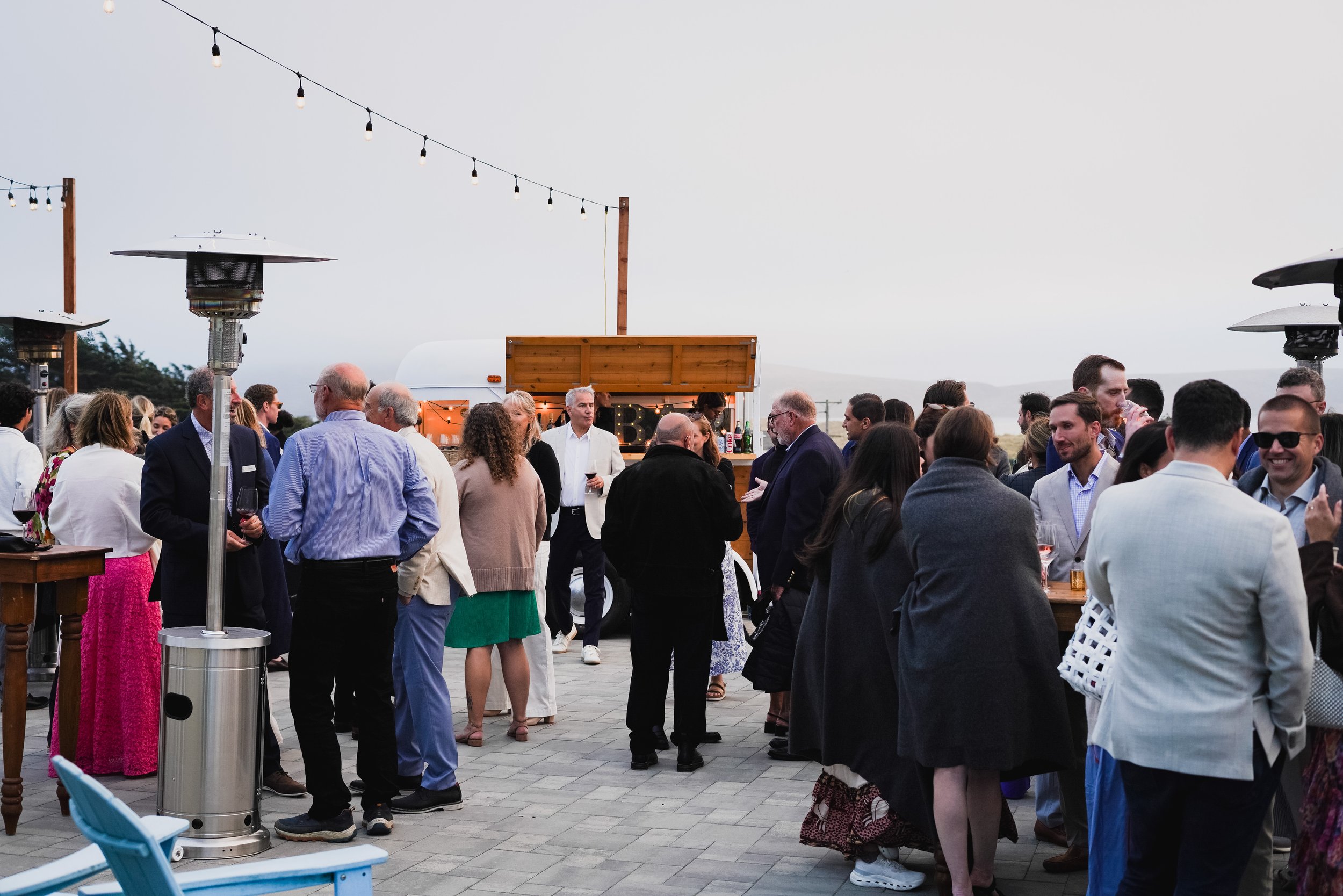 An outdoor gathering with people mingling near a bar and patio heaters, under string lights with a cloudy sky in the background.