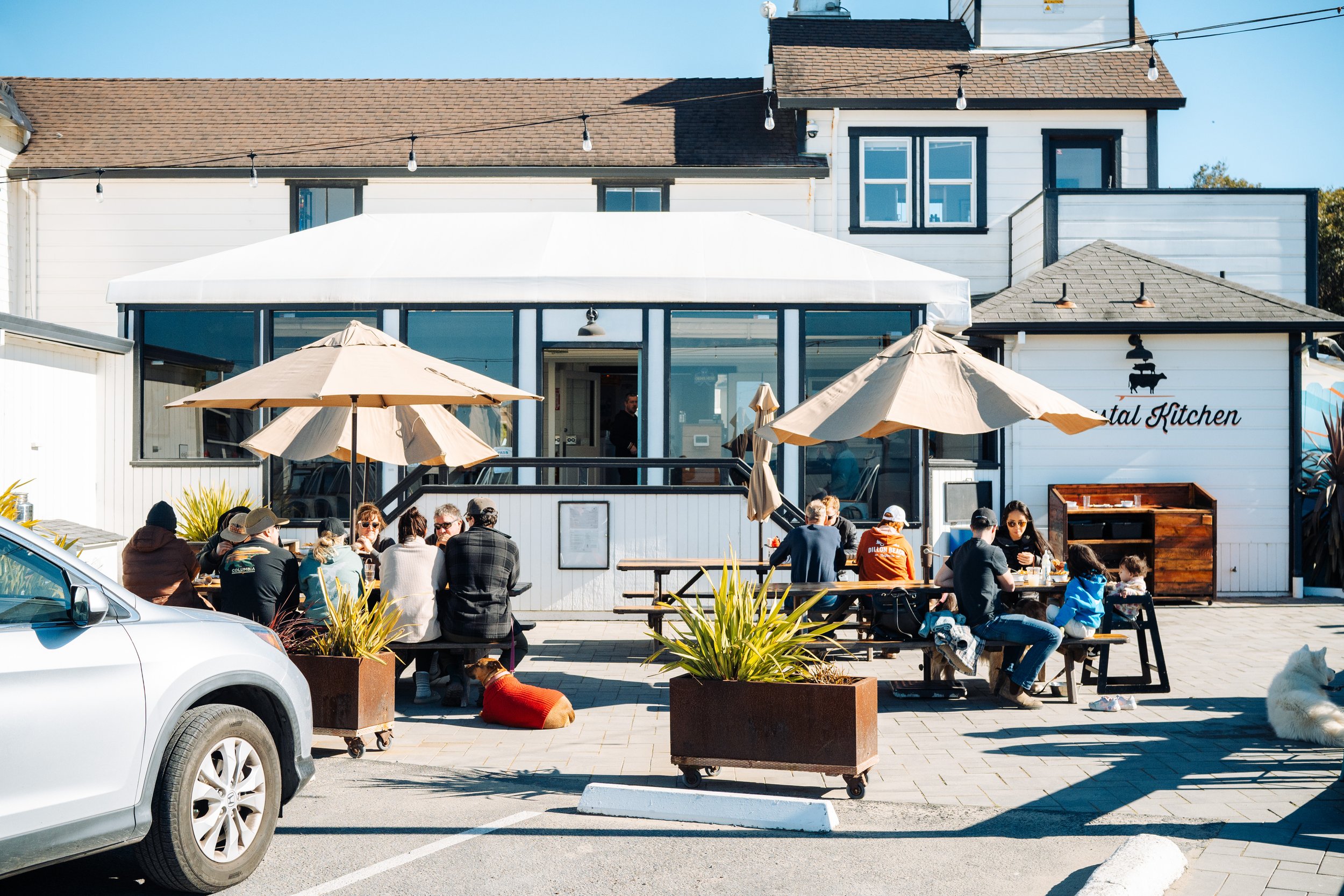 People dining outdoors at a restaurant with umbrellas, with a building in the background that has a sign reading 'Rental Kitchen' and a dog resting on the ground.