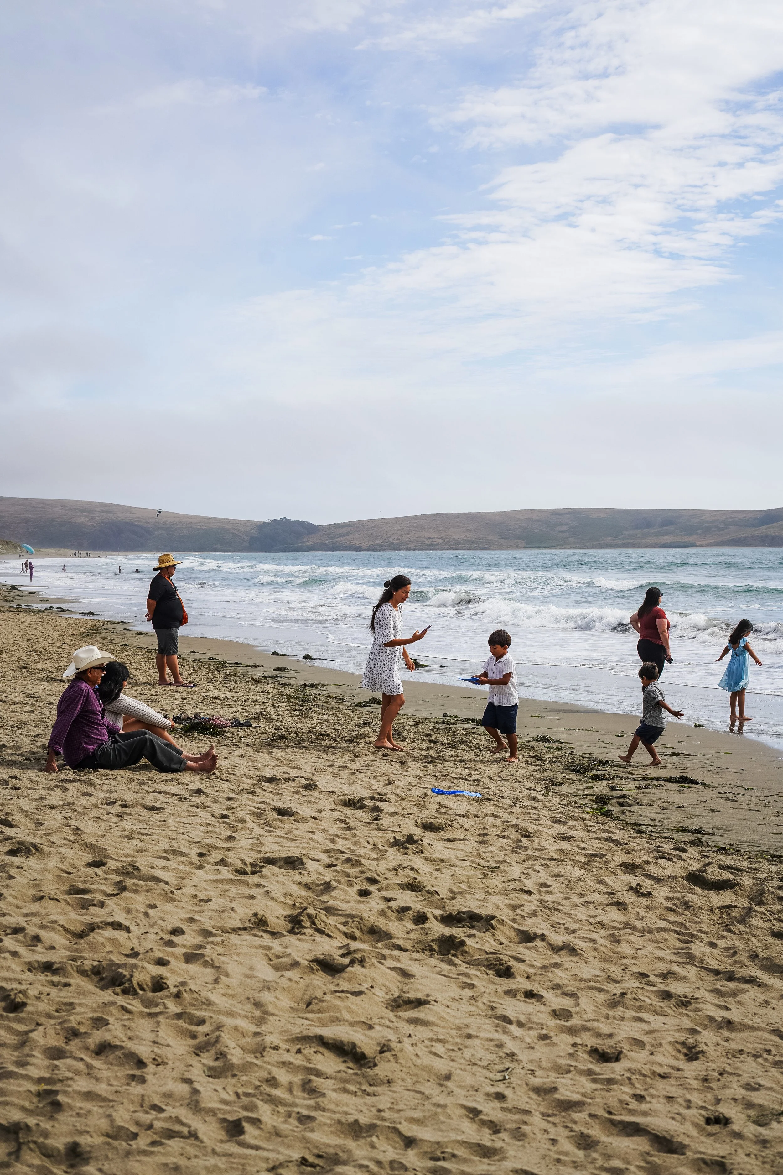 People enjoying a day at the beach; some are sitting on the sand, others are standing or walking near the shoreline with small waves. The sky is partly cloudy with a distant hilly landscape in the background.