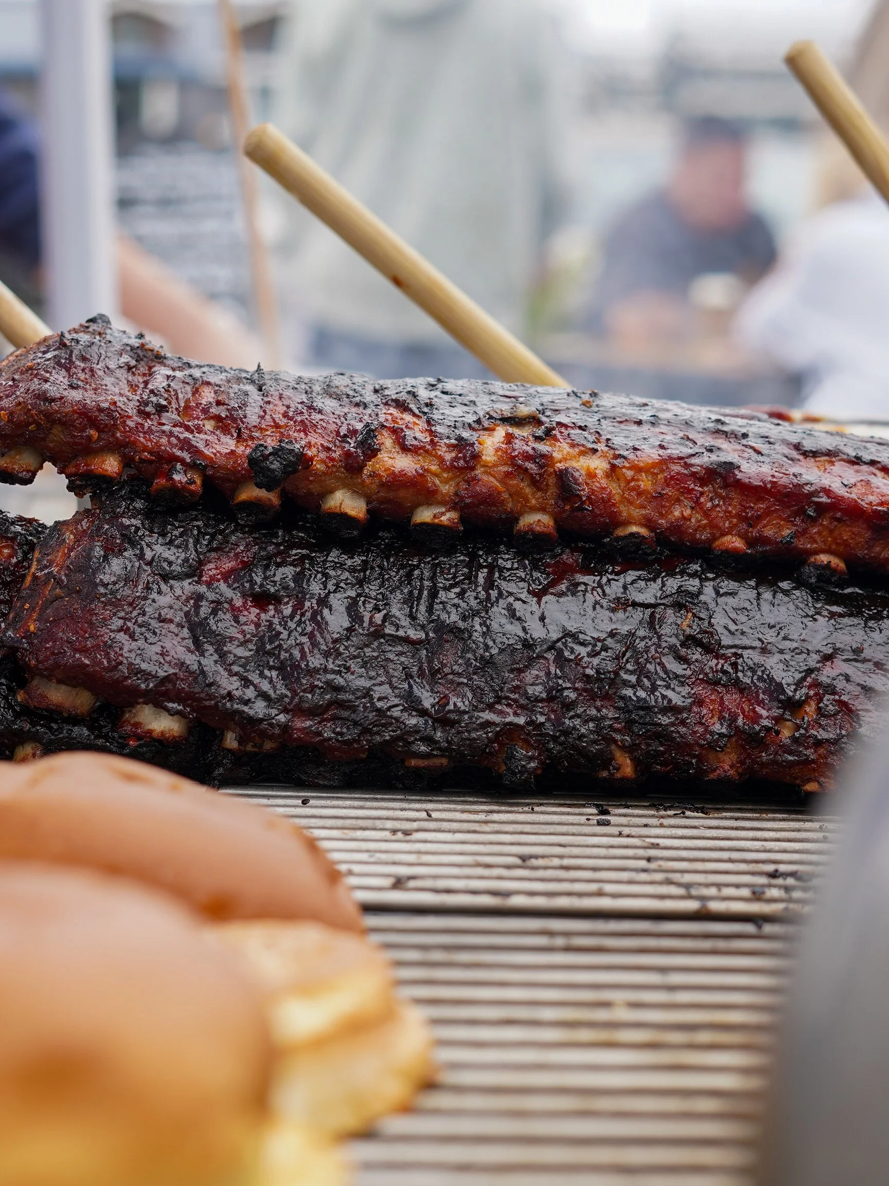 Close-up of a rack of barbecue ribs with a glaze, with some blurred hot dogs in the foreground. In the background, a person is slightly blurred, standing behind a smoky outdoor grill or cooking area.