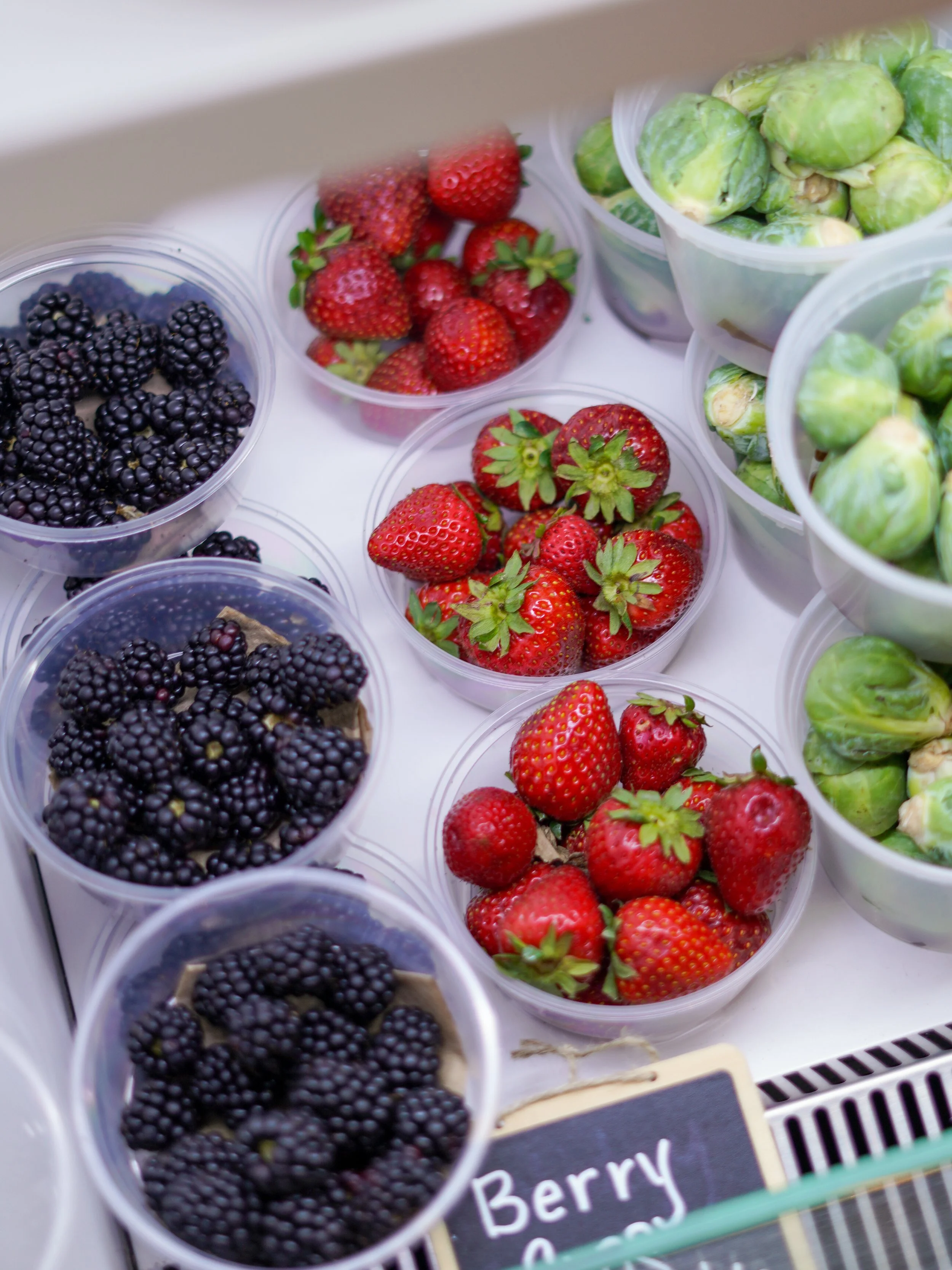 Plastic containers of fresh berries including blackberries, strawberries, and green berries at a market or grocery store.