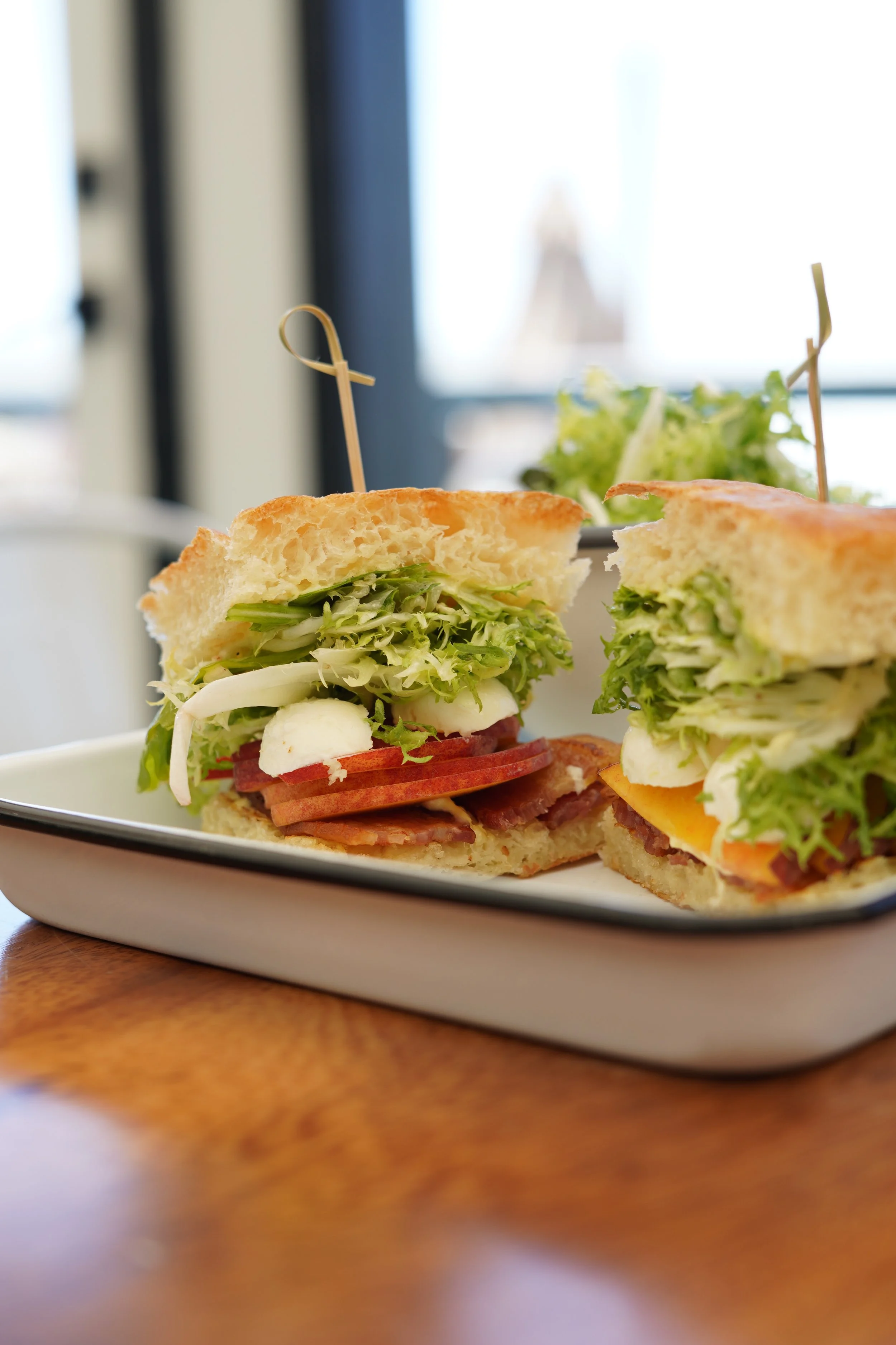 Close-up image of a sandwich on a white rectangular plate placed on a wooden table, with a blurred background of a window and greenery.