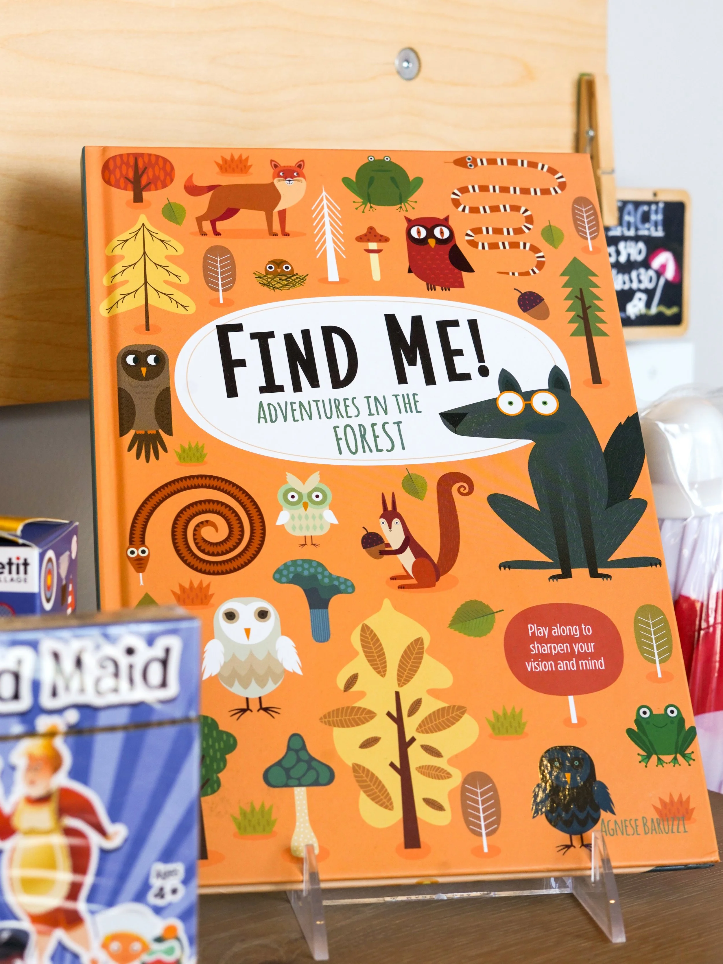 A colorful children's book titled "Find Me! Adventures in the Forest" on display, featuring illustrations of animals like a fox, frog, owl, snake, and owl, along with trees and leaves, with a small chalkboard in the background.