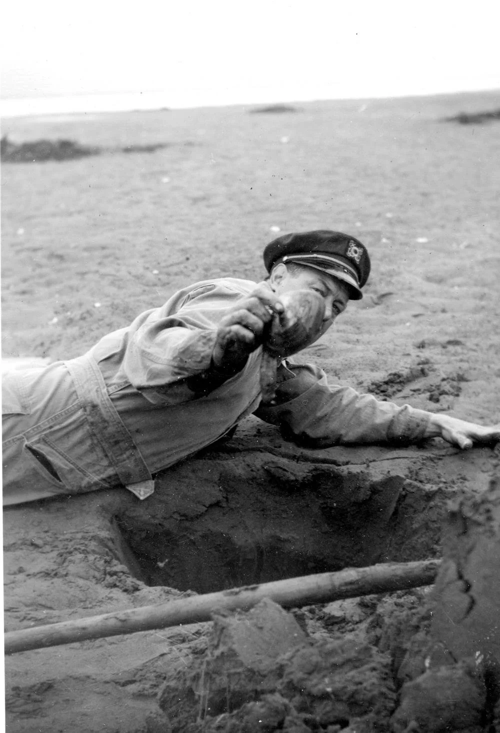A black-and-white photo of a man in a captain's hat and coveralls lying on a beach, holding up a large Pacific razor clam.