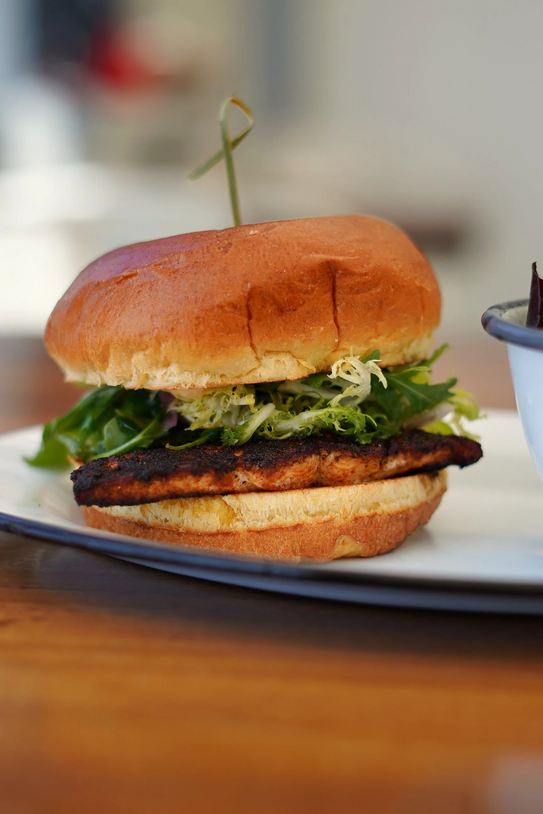 Close-up of a sandwich with a toasted bun, lettuce, a grilled patty, and other sandwich ingredients on a white plate on a wooden table.