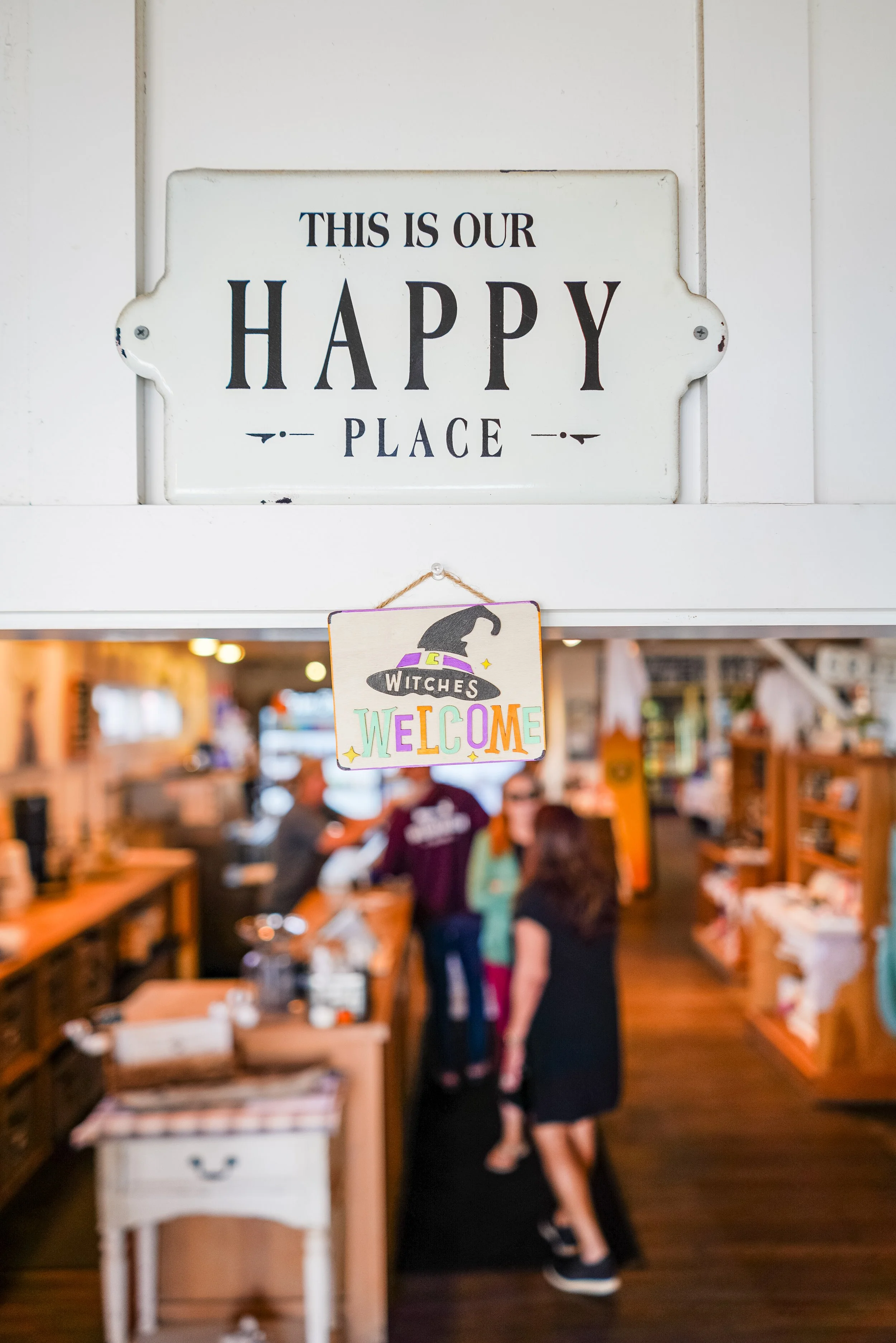 A white sign with black text that reads "This is our happy place" is mounted on a wooden wall. Below the sign, a colorful Halloween-themed hanging decoration says "Witches Welcome." In the background, there are several people inside a cozy, well-lit store or café, with wooden furniture and warm lighting.