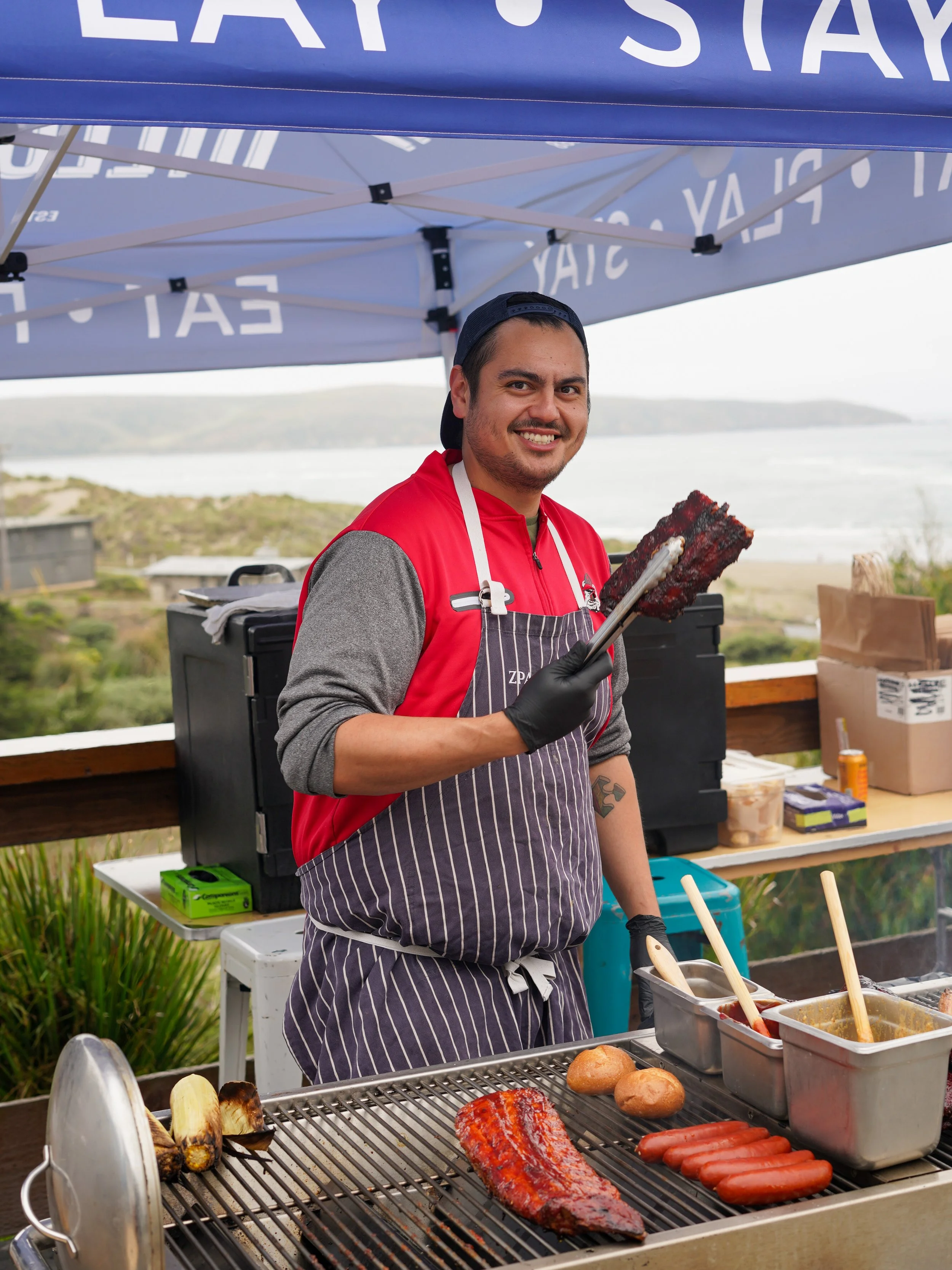 Man grilling meats on an outdoor barbecue at a cookout event, with various food items and grilling accessories on the grill and table.