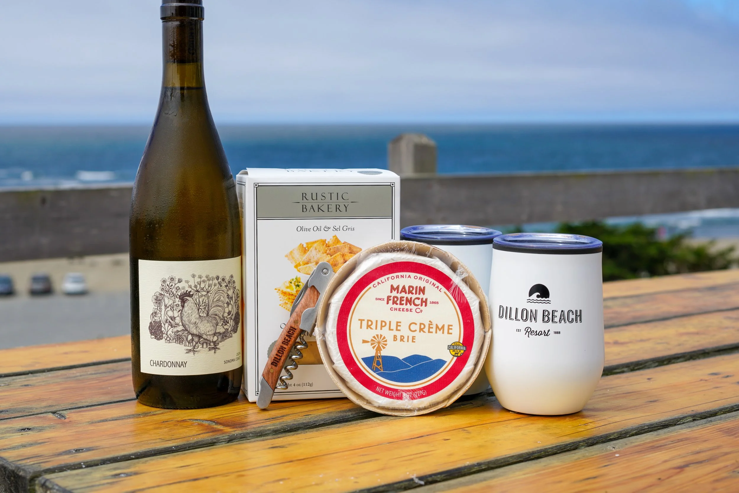 A bottle of Chardonnay wine, a box of Rustic Bakery olive oil and sea salt crackers, a package of Marin French triple crème brie cheese, and two white cups, with a cloudy sky and ocean in the background, on a weathered wooden table.