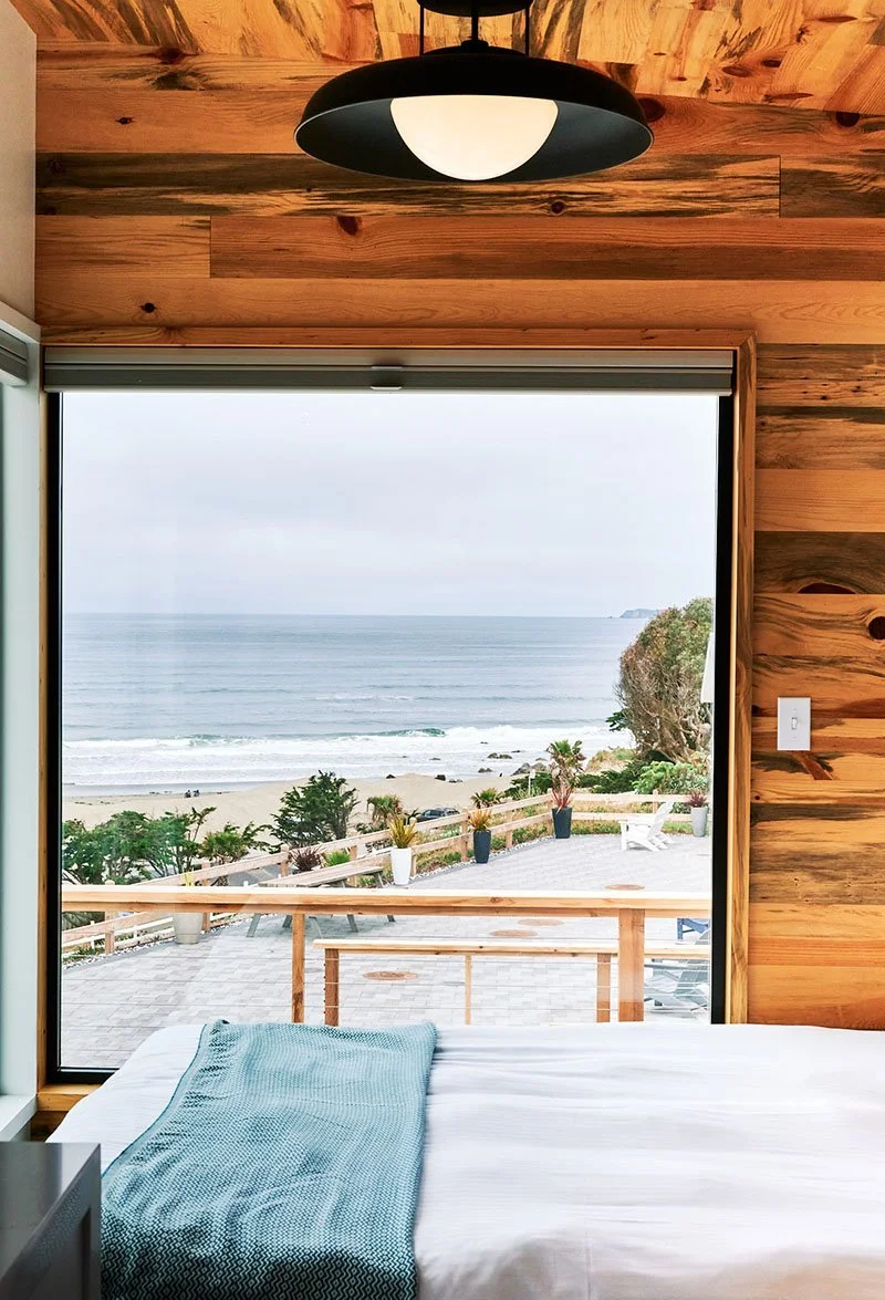 Interior of a modern Osprey cabin at Dillon Beach Resort featuring a gray sofa with a yellow textured pillow, mid-century modern coffee table, and large floor-to-ceiling windows overlooking grassy dunes and the ocean.