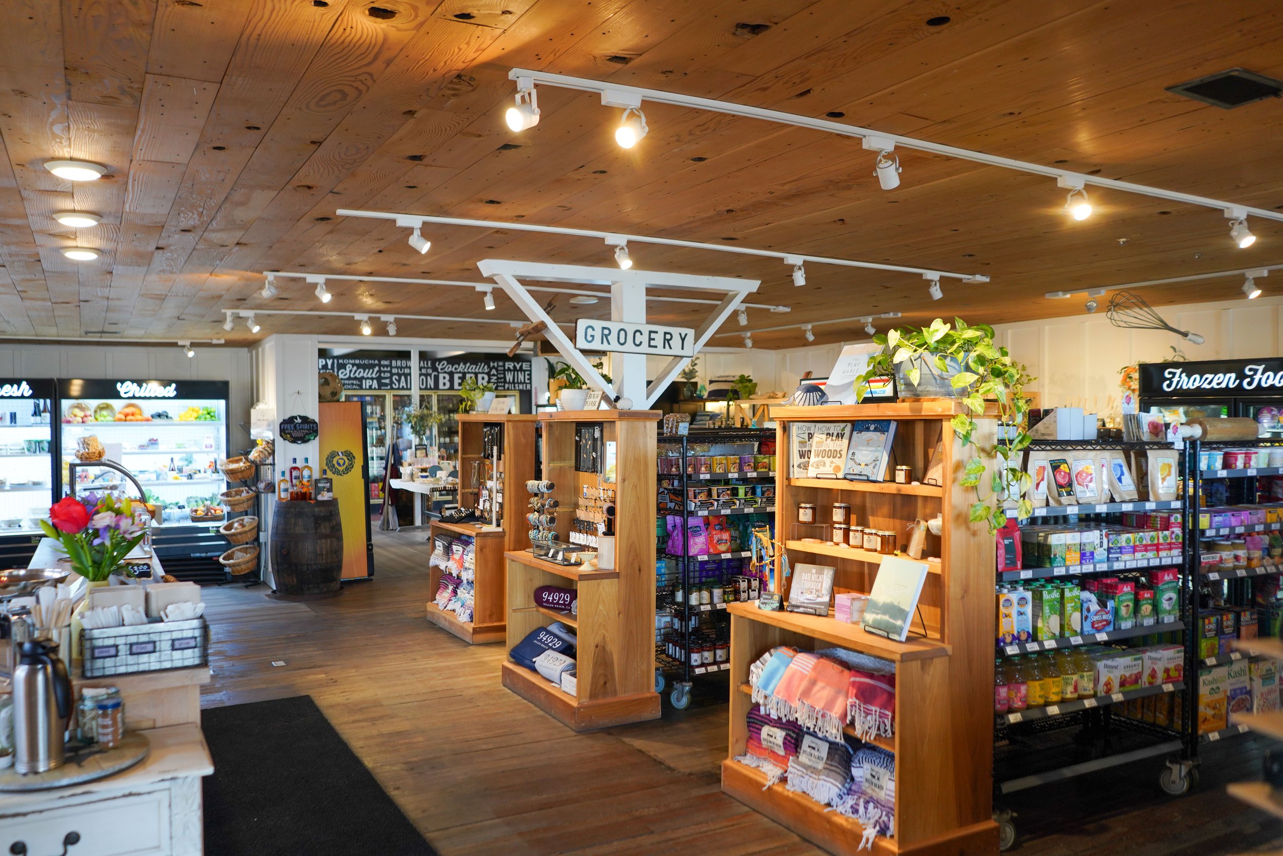 Interior view of a grocery store with wooden ceiling and various shelves stocked with food and household items, including beverages, snacks, and condiments.