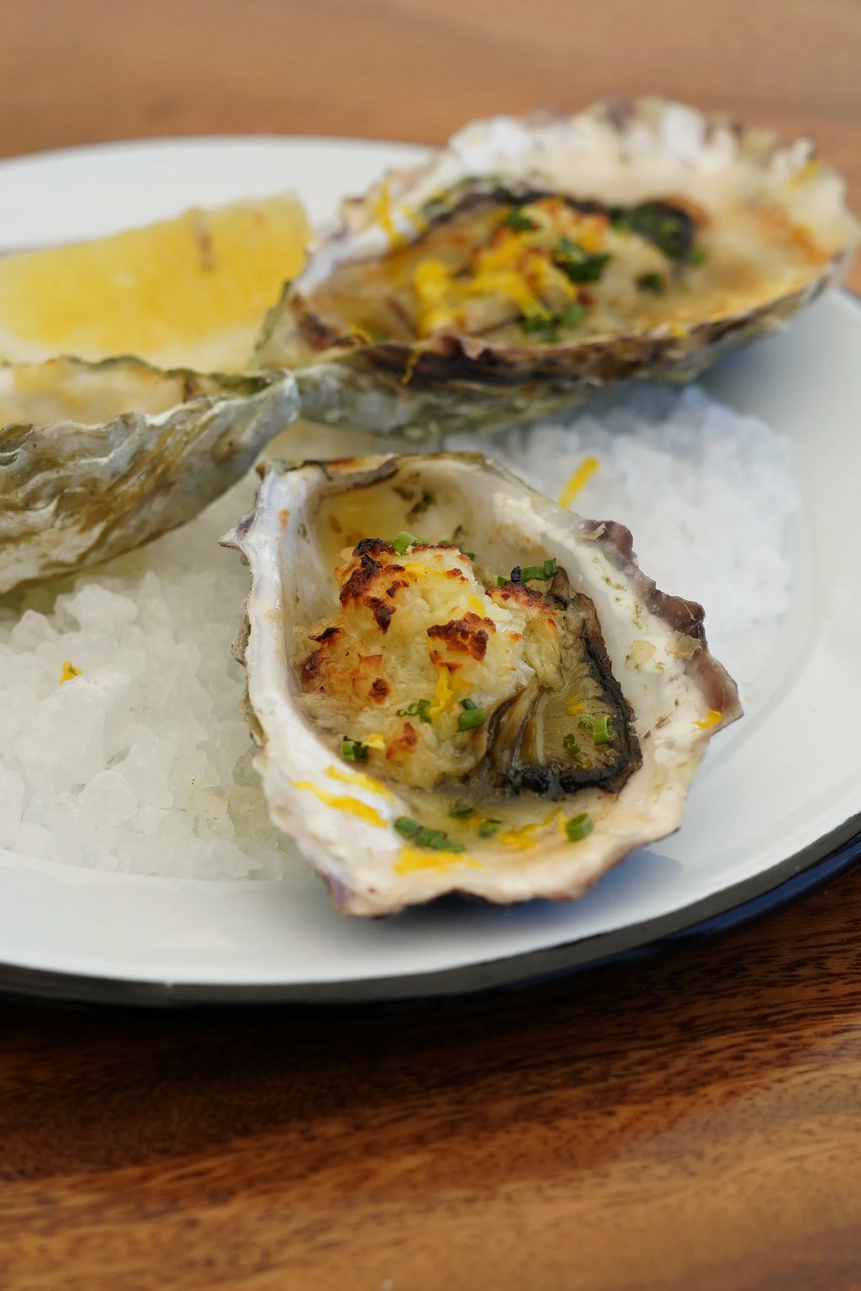 A white plate with ice, two oysters on the half shell garnished with herbs, and a lemon wedge on a wooden table.