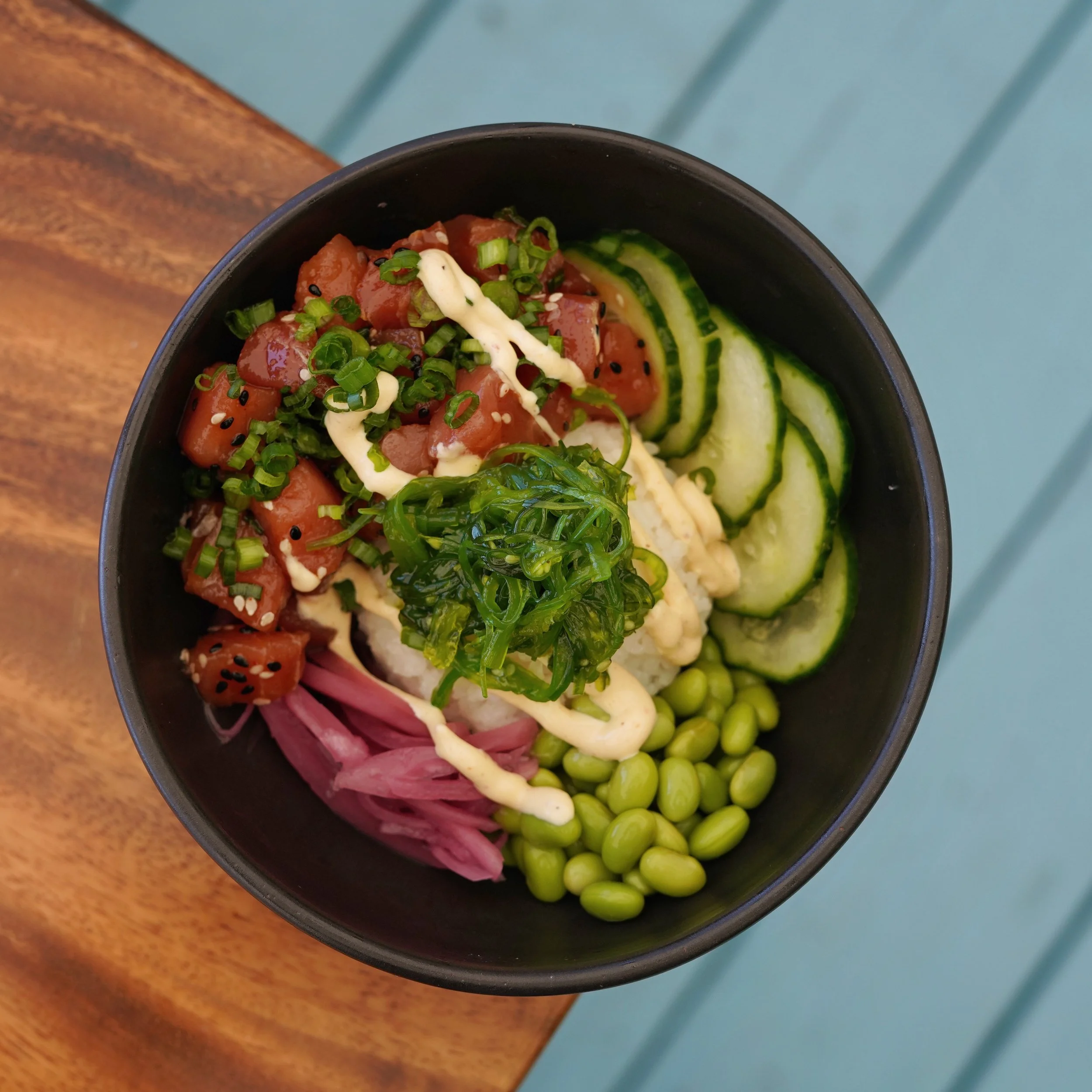A black bowl filled with chopped raw tuna, sliced cucumbers, seaweed salad, and garnished with chopped green onions, drizzled with a creamy sauce and sprinkled with sesame seeds. The bowl is placed on a wooden table near a teal-colored surface.