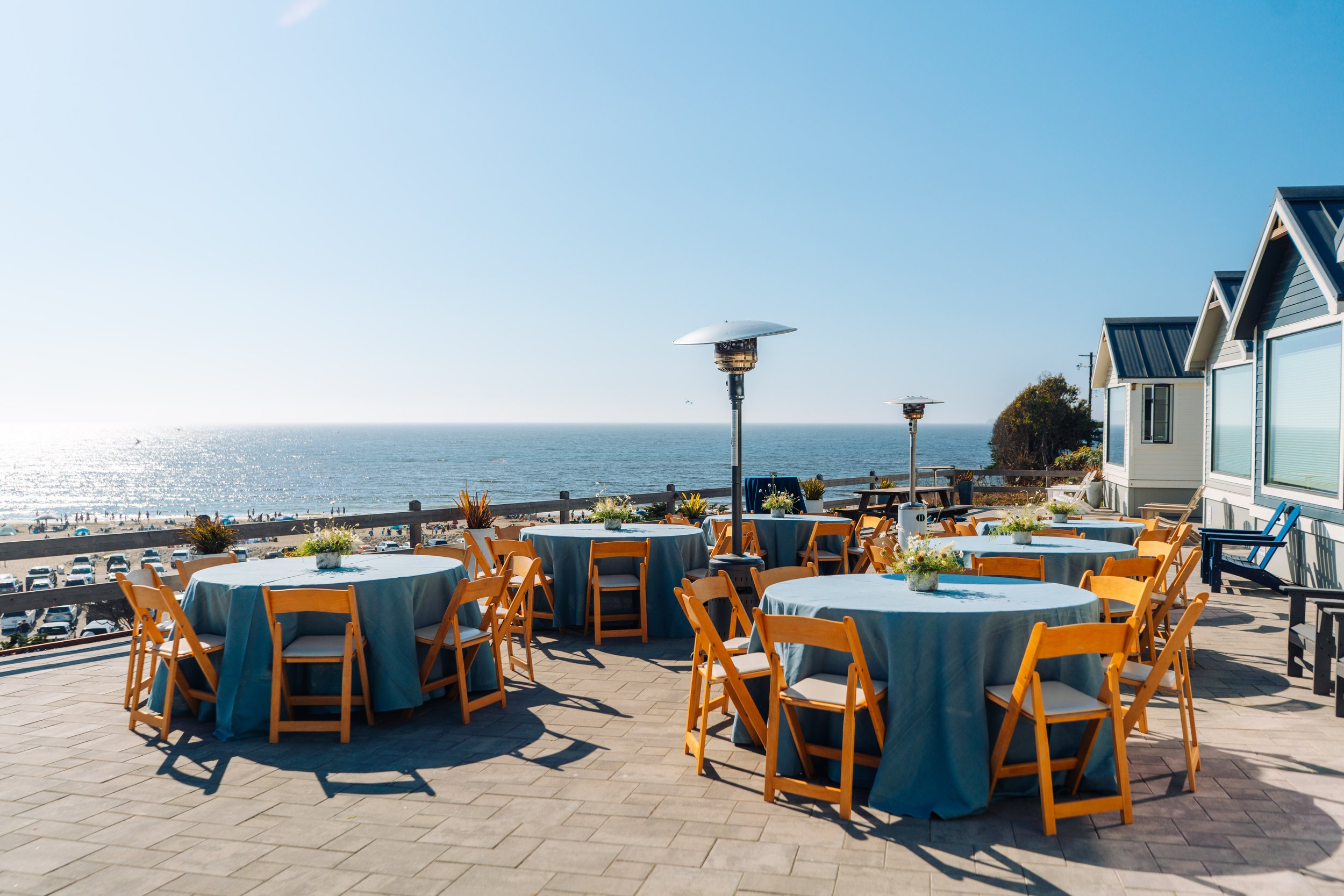 Outdoor restaurant patio overlooking a beach with tables covered in blue tablecloths and yellow chairs, with the ocean and sky in the background.
