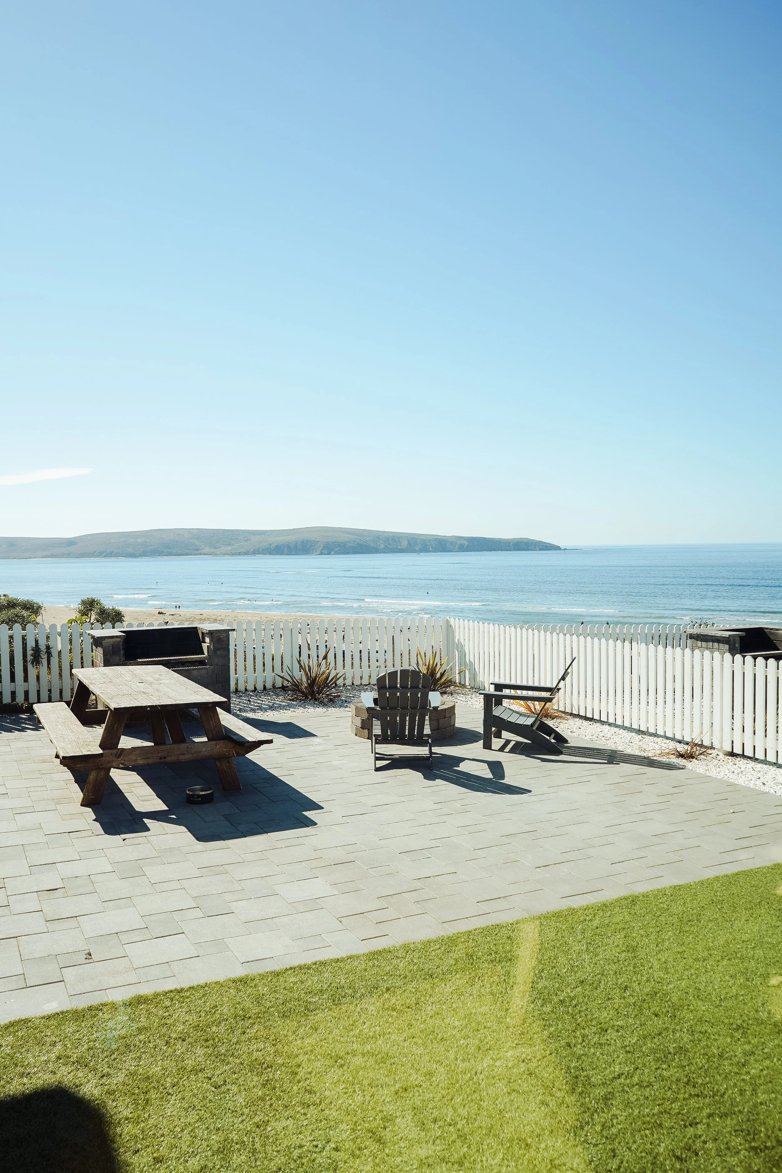 Stone paver patio with a wooden picnic table, Adirondack chairs, and a fire pit overlooking a sandy beach and ocean under a clear blue sky.