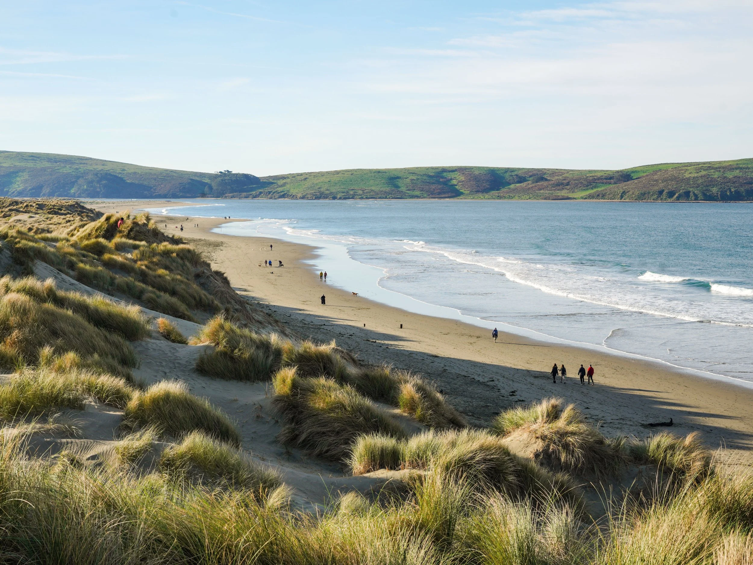 A sandy beach with green grassy dunes in the foreground. Several people are walking along the shoreline near the water, and a few are walking on the dunes. The ocean is calm with gentle waves, and green hills are visible in the background under a cle