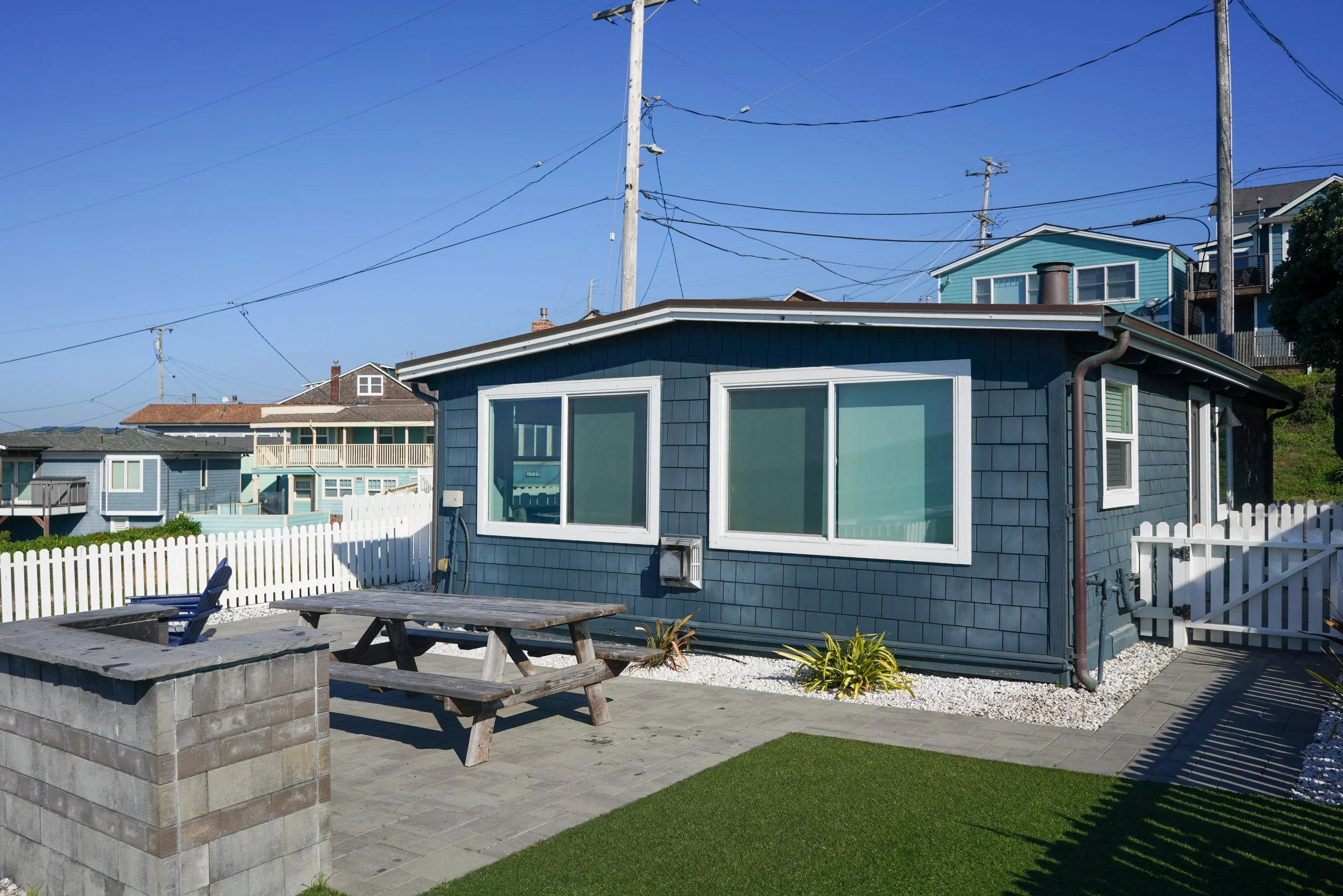 A detailed view of a private backyard patio with a modern concrete block fire pit, a weathered wooden picnic table, and blue shingle siding on the house, all enclosed by a white picket fence.
