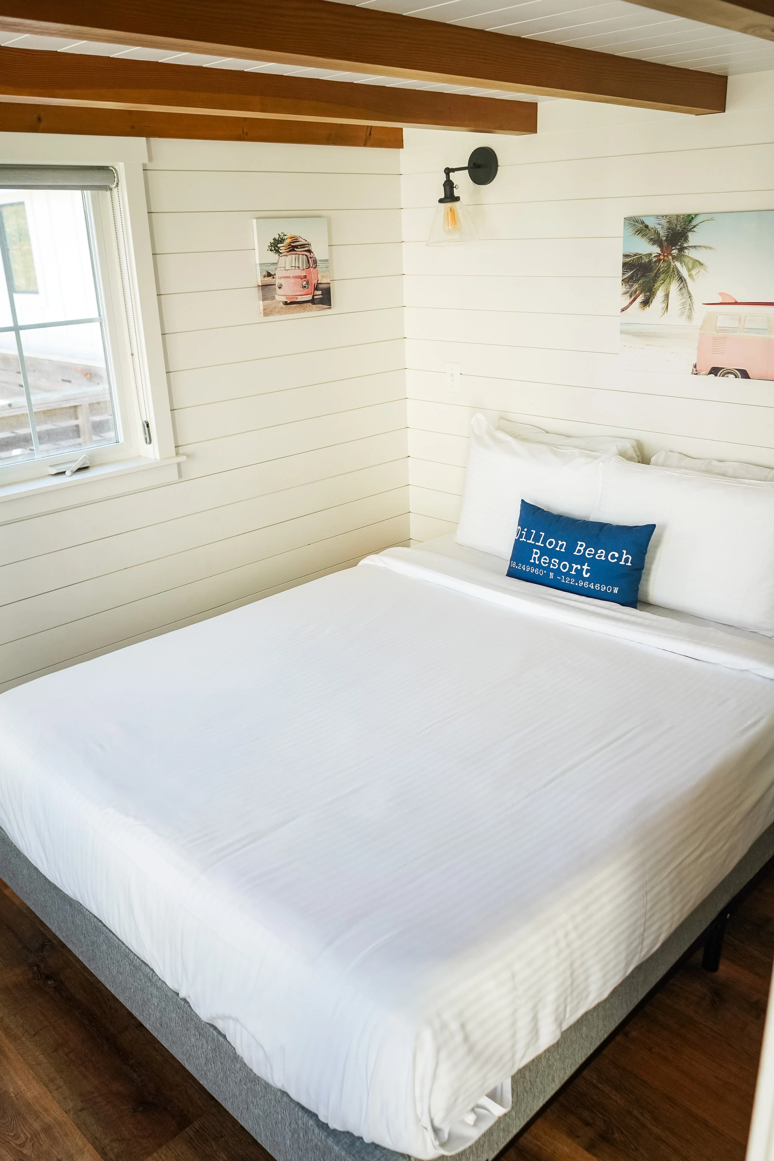 A neatly made white bed with a blue pillow that says 'Dillon Beach Resort' in a room with shiplap walls, two wall-mounted light fixtures, and artwork featuring a vintage pink van, surfboard, palm trees, and a beach scene.