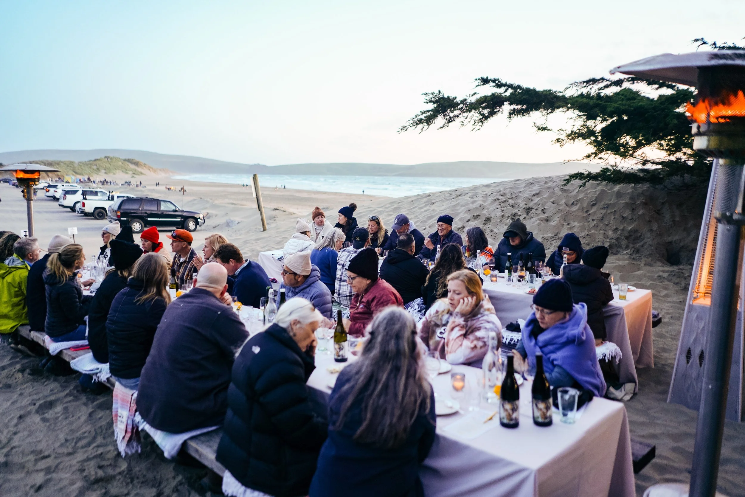 People enjoying an outdoor dinner on a beach at sunset, with tables set up on the sand and cars parked nearby, and heaters providing warmth.