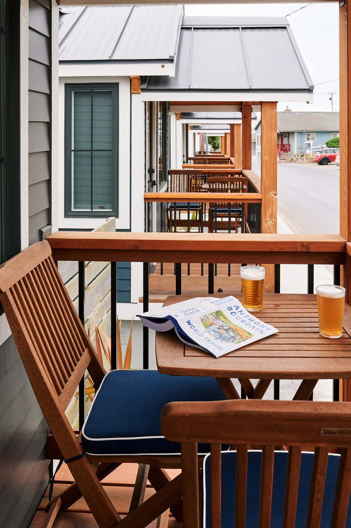 Private wooden deck of a Sandpiper cabin featuring a bistro table with two beers and an Anthony Bourdain book, overlooking a row of similar modern cottages.