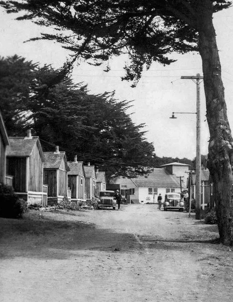 A vintage B&W photo of a dirt road lined with rustic wood cabins, two mid-century cars, and people near a larger white building.