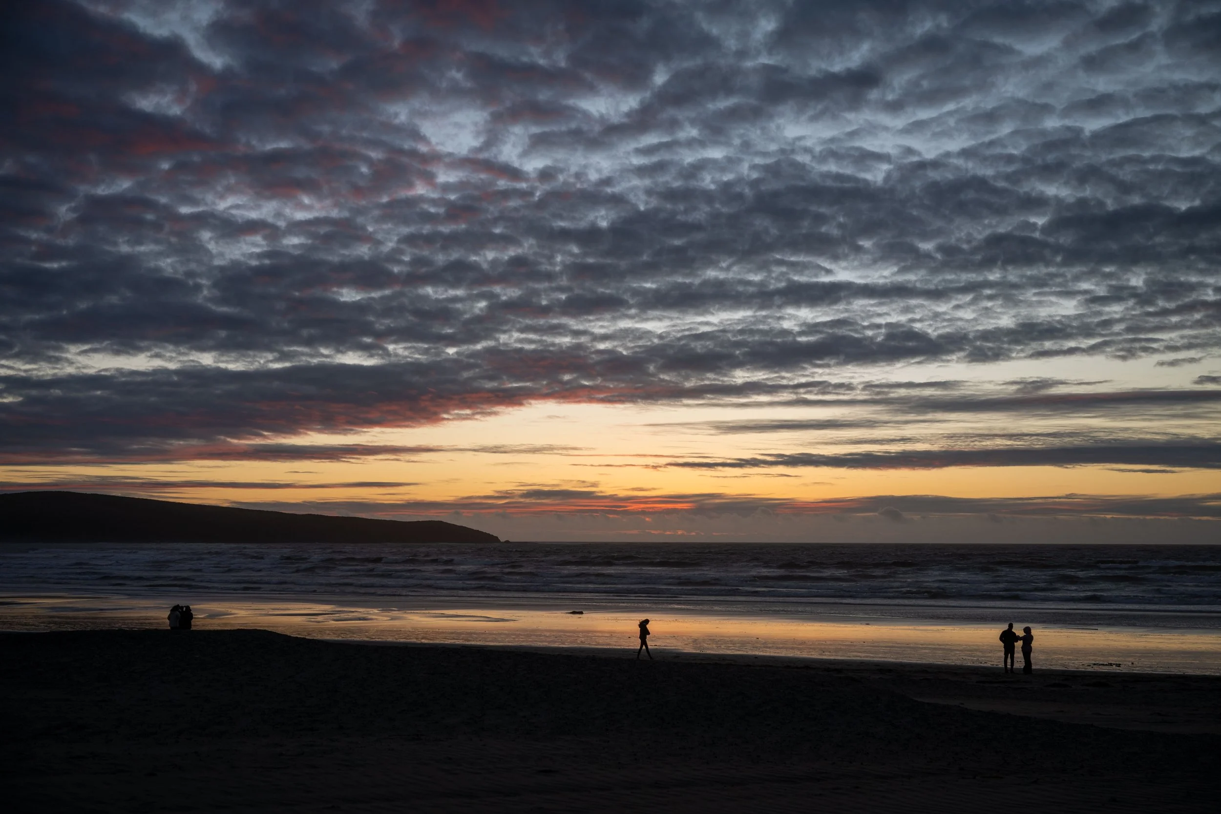 A sunset over the ocean with a dark silhouette of people on the beach, some walking and some standing, with a sky filled with layered clouds.