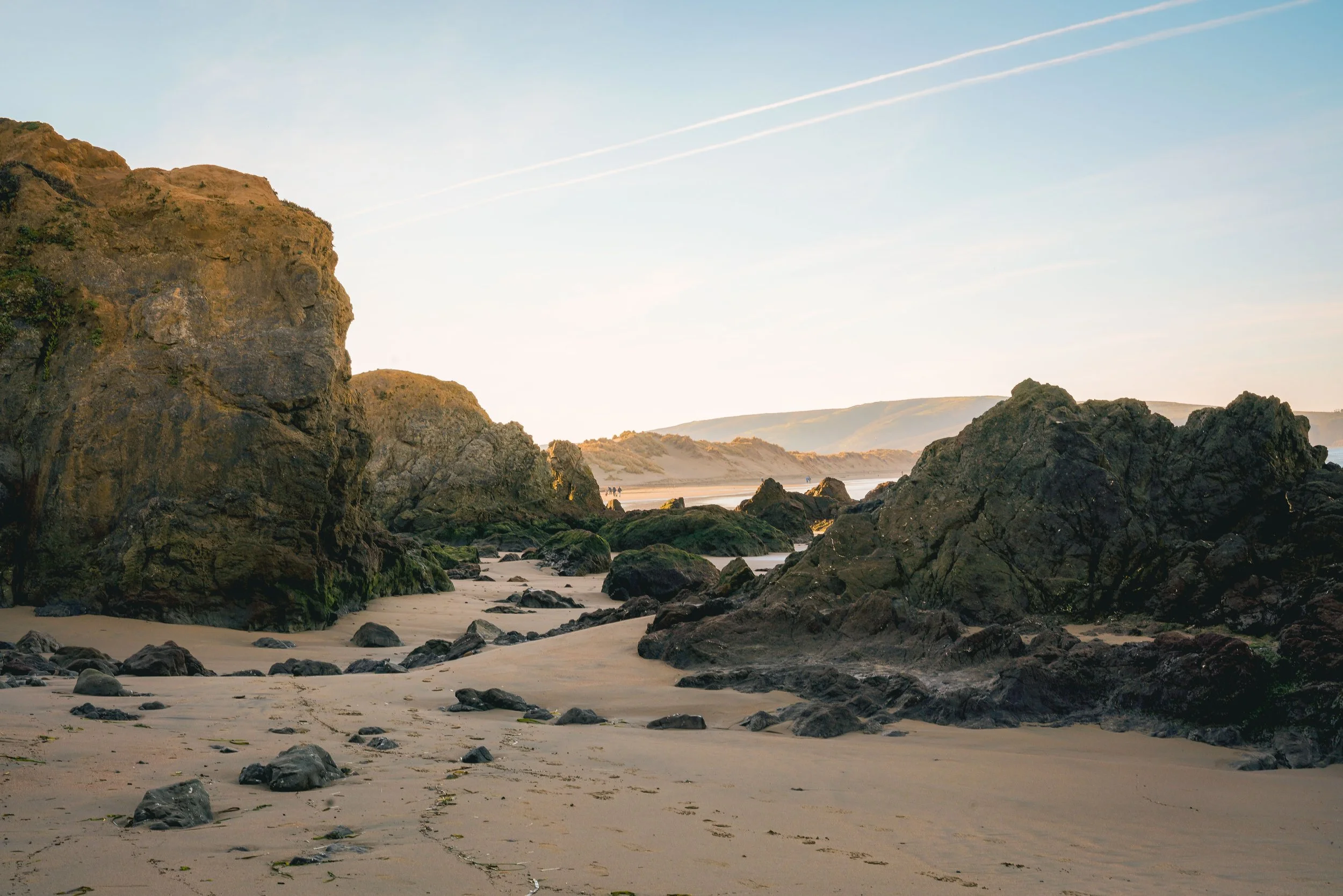 Rocky coastline with large boulders and green moss, sandy beach, distant hills, and a clear sky with a few contrails.