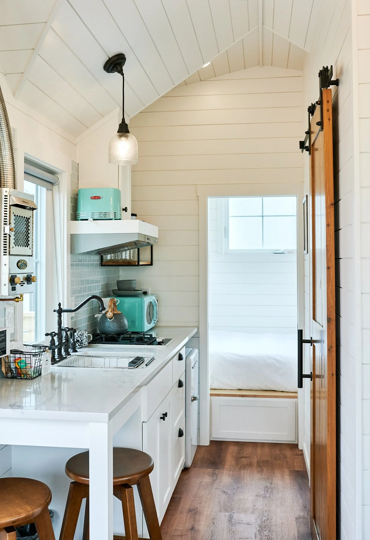 A small, modern kitchen with white cabinets, a white countertop, a small sink, a gas stove, and a microwave. There are two wooden stools at a white counter, and a hanging light fixture. The kitchen has white shiplap walls and ceiling, and a sliding b