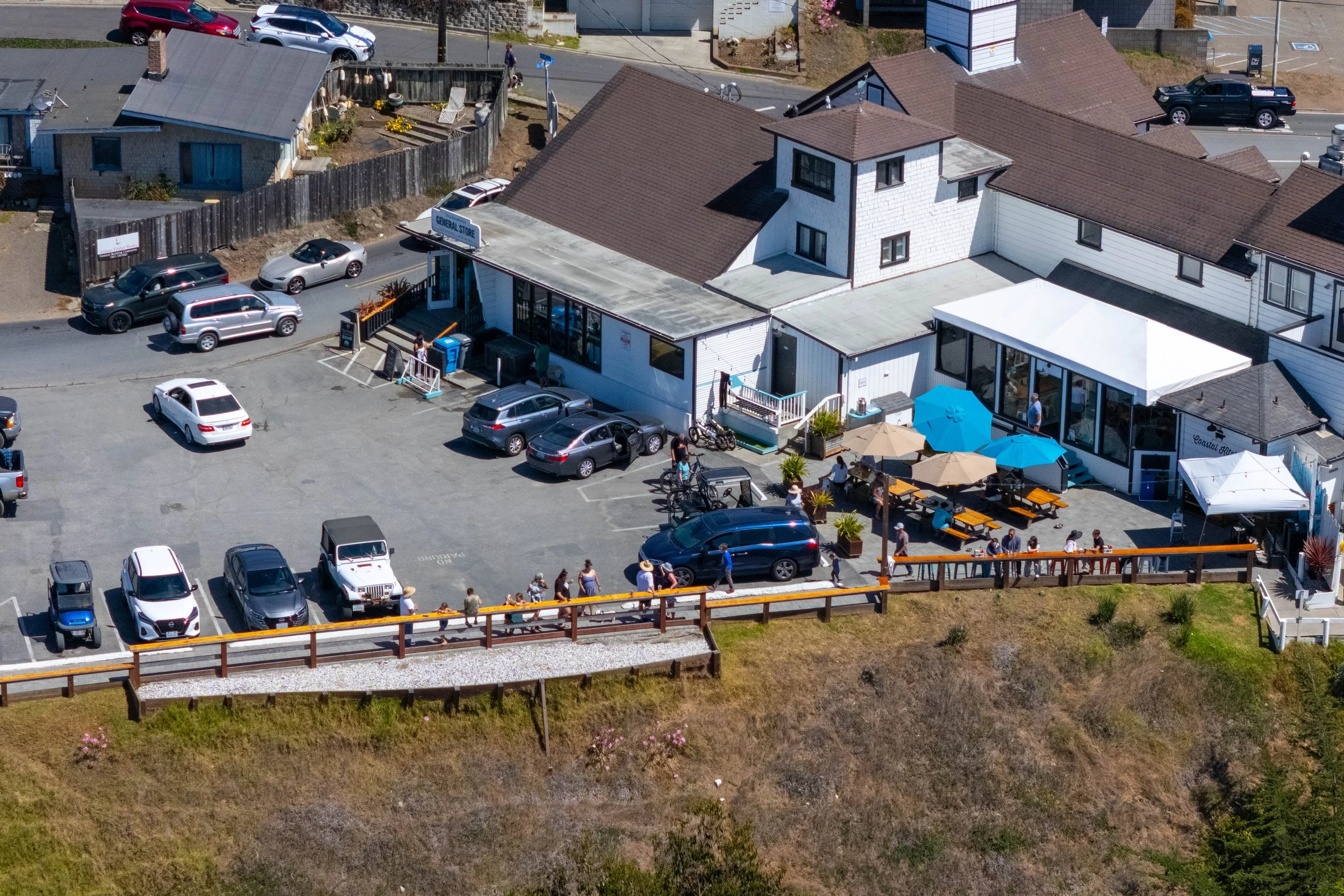 An aerial view of a small coastal town or village with a parking lot filled with cars, a building with outdoor seating shaded by umbrellas, and surrounding residential houses. People are seen walking and gathering outside, and some are sitting at tab